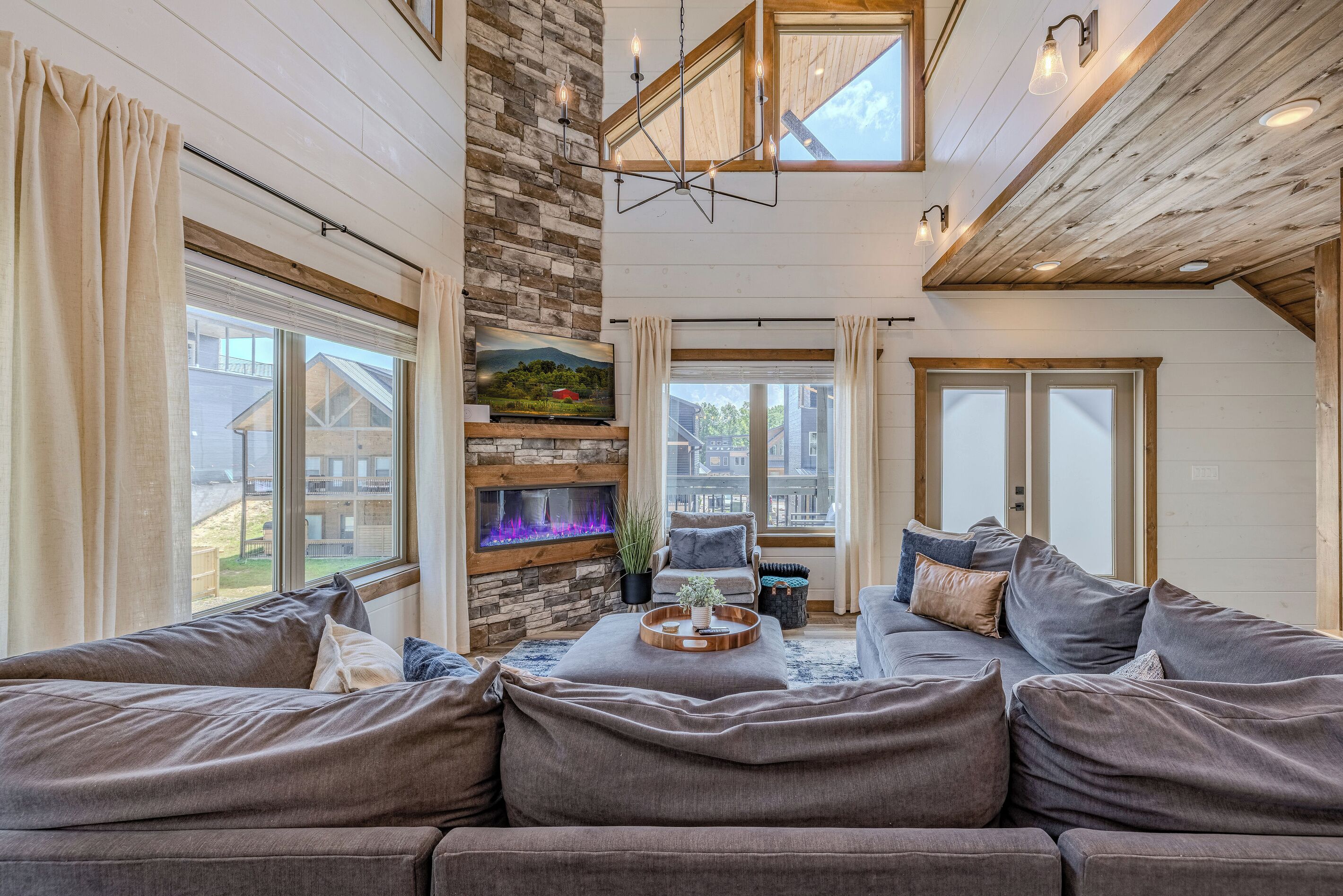A cozy living room with a stone fireplace, a large window, and a high ceiling with exposed beams.