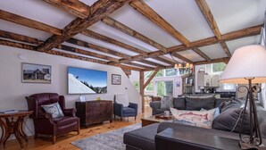 A cozy living room with wooden beams, a fireplace, a sofa, a coffee table, and a dining area with a kitchen in the background.