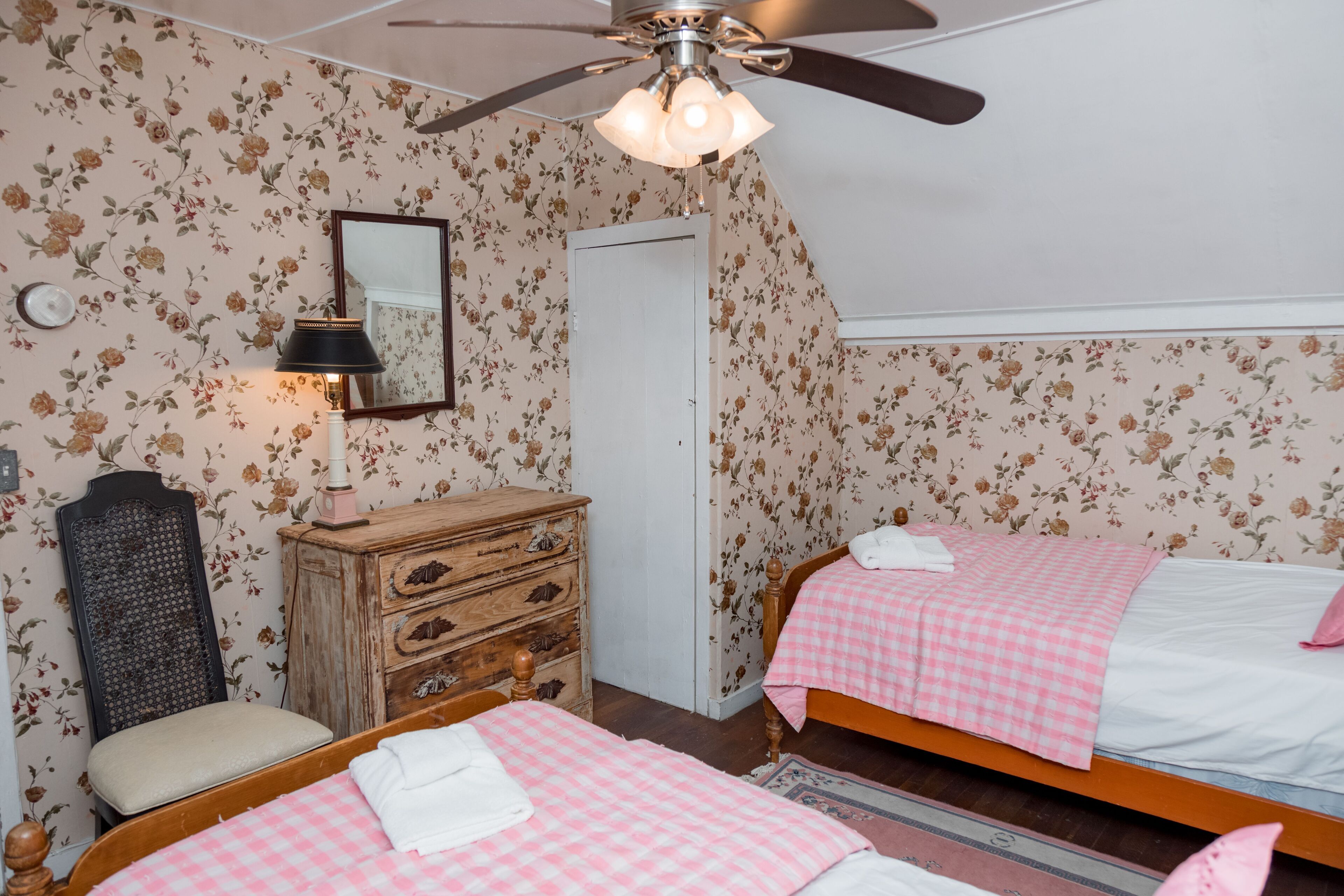 A bedroom with floral wallpaper, a ceiling fan, a mirror, a wooden dresser, and two beds with pink checkered bedding.