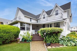 A two-story house with a front porch, a covered entryway, and a small front yard with a gravel path leading to the door. - Historic Oak Bluffs Cottage – Porch with View (Oak Bluffs)