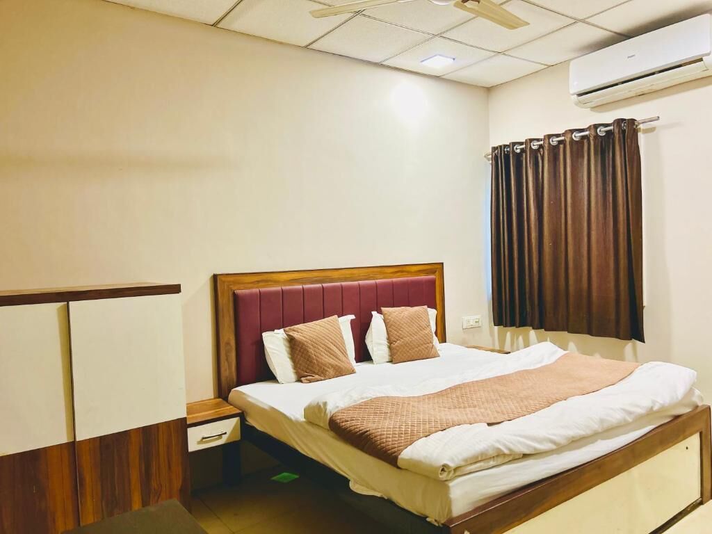 Una habitación de hotel con una cama, una mesita de noche, un cabecero de madera, una ventana con cortinas y un equipo de aire acondicionado.
