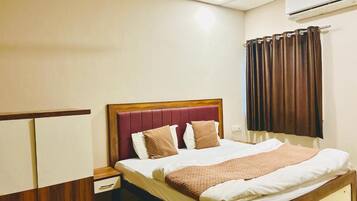 Una habitación de hotel con una cama, una mesita de noche, un cabecero de madera, una ventana con cortinas y un equipo de aire acondicionado.
