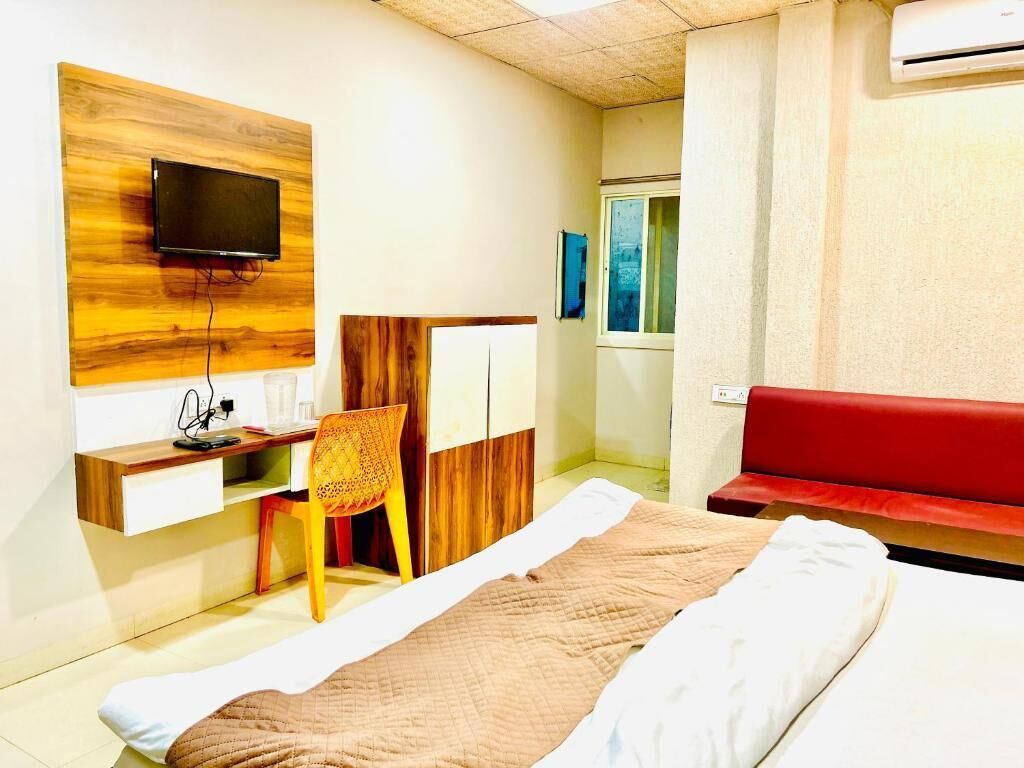 A hotel room with a bed, a desk with a computer, a television mounted on a wooden panel, a red sofa, and an air conditioning unit.