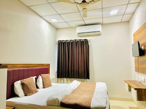 A hotel room with a bed, a ceiling fan, an air conditioning unit, a TV mounted on the wall, and a wooden side table. - Hotel Jagtara (Indore)