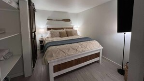 Un dormitorio con cama, mesitas de noche, lámpara, luz de techo y televisor montado en la pared.
