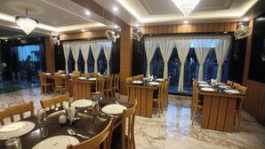 A dining area with marble flooring, wooden tables, and chairs. There are chandeliers and large windows with curtains.