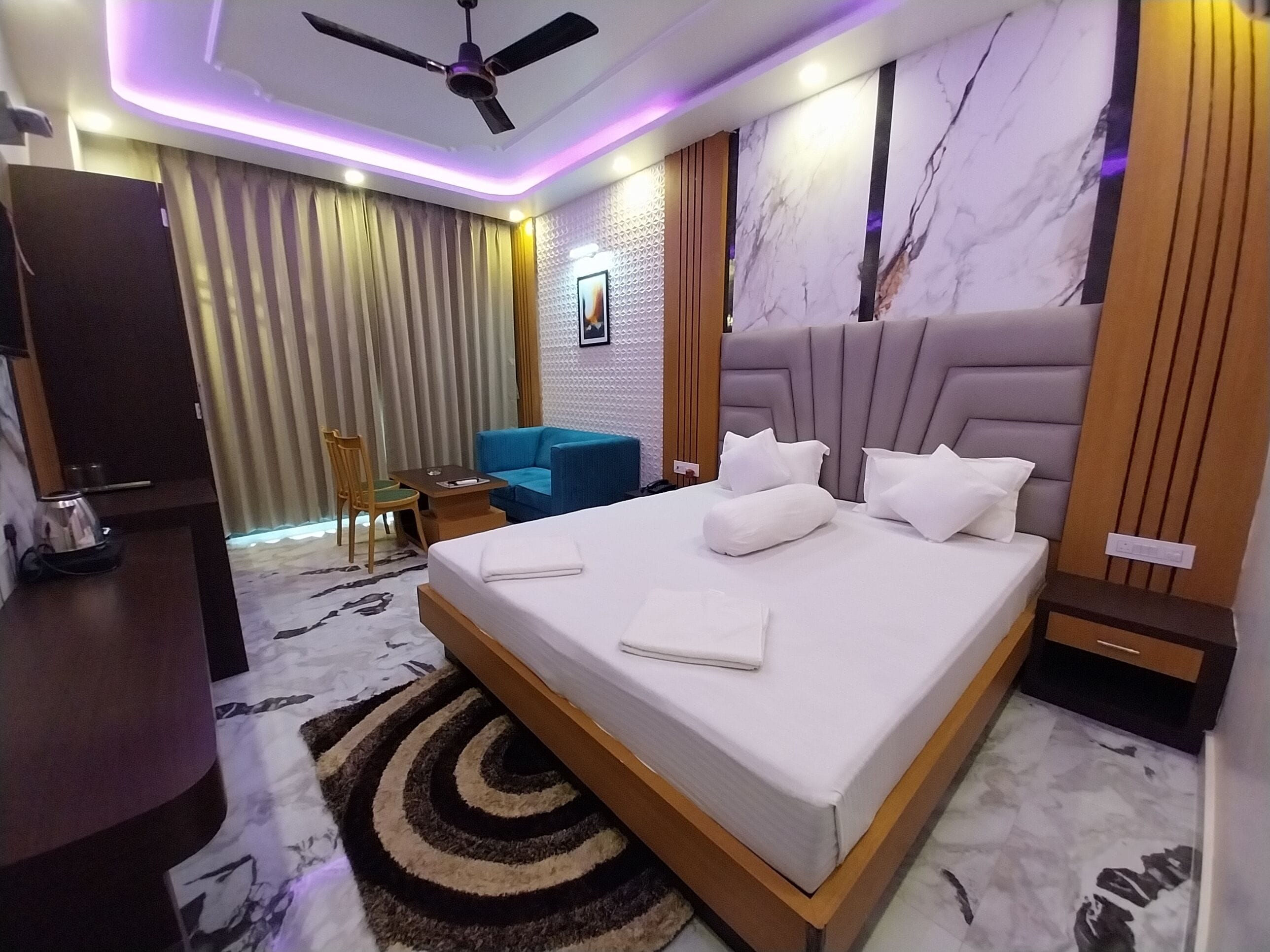 Ein modernes Hotelzimmer mit einem großen Bett, einem Deckenventilator, einem blauen Sessel, einem kleinen Tisch und einem Fernseher.