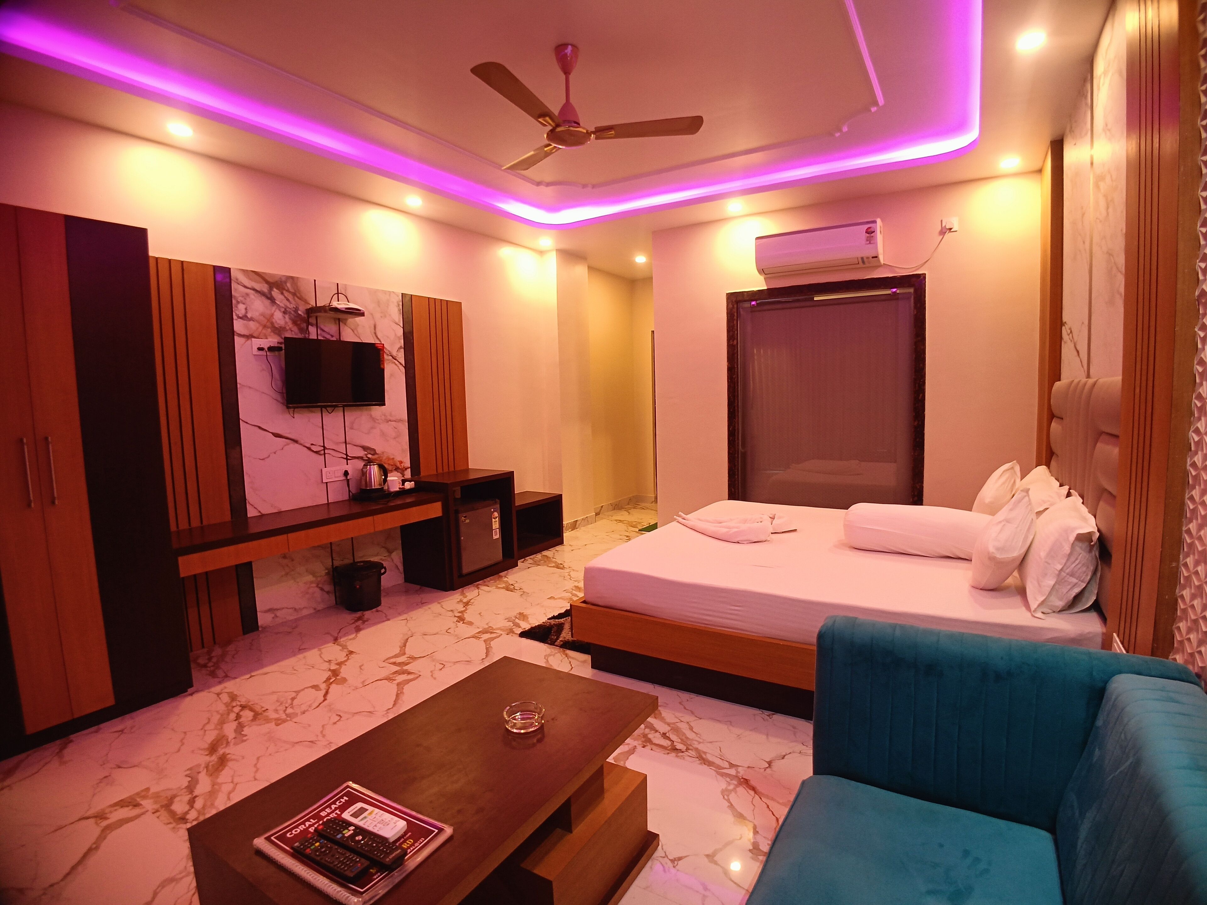 A modern hotel room with a bed, a blue armchair, a wooden coffee table, a TV, and a ceiling fan.