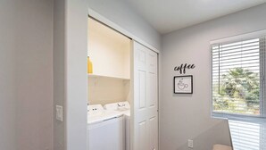 A laundry room with a white door, a washing machine, and a dryer. There is a window with blinds and a wall-mounted picture with the word "coffee".