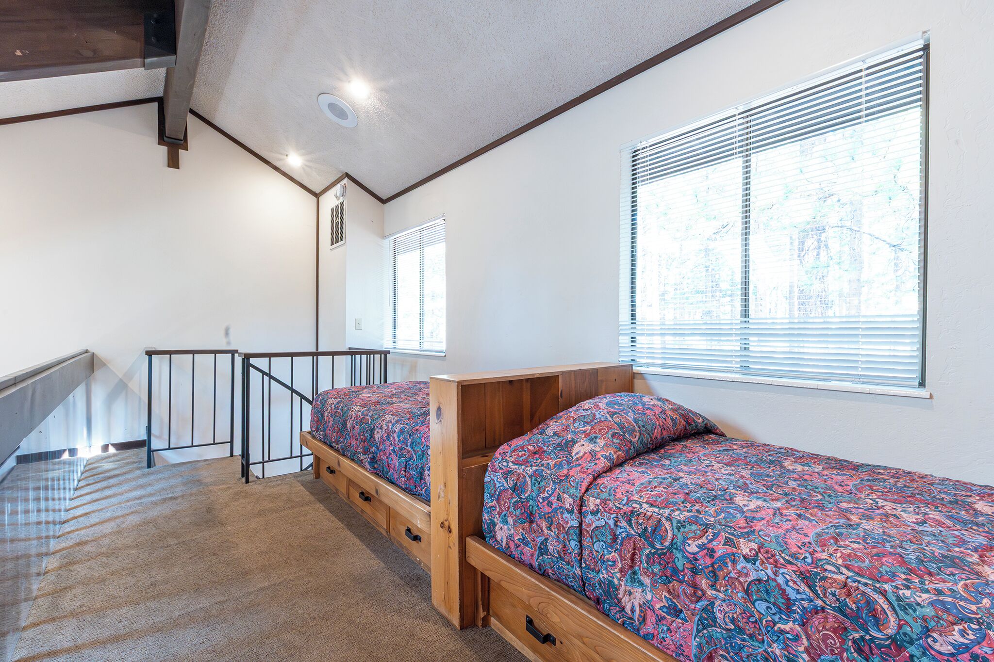 Une chambre avec deux lits simples, une tête de lit en bois, un sol recouvert de moquette, un escalier et une fenêtre avec des stores.