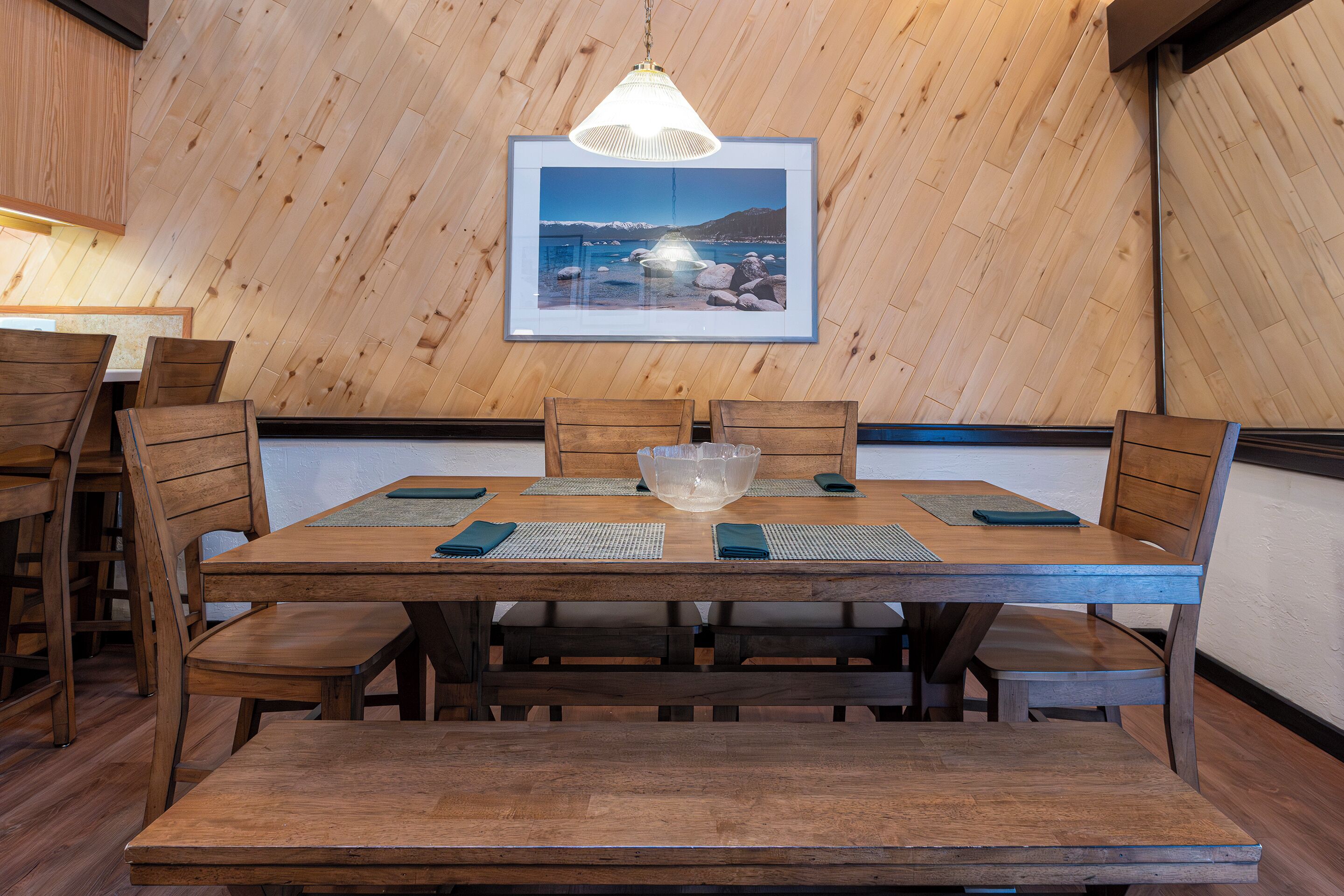 Une table à manger en bois avec des bancs, un luminaire suspendu et un tableau encadré au mur.