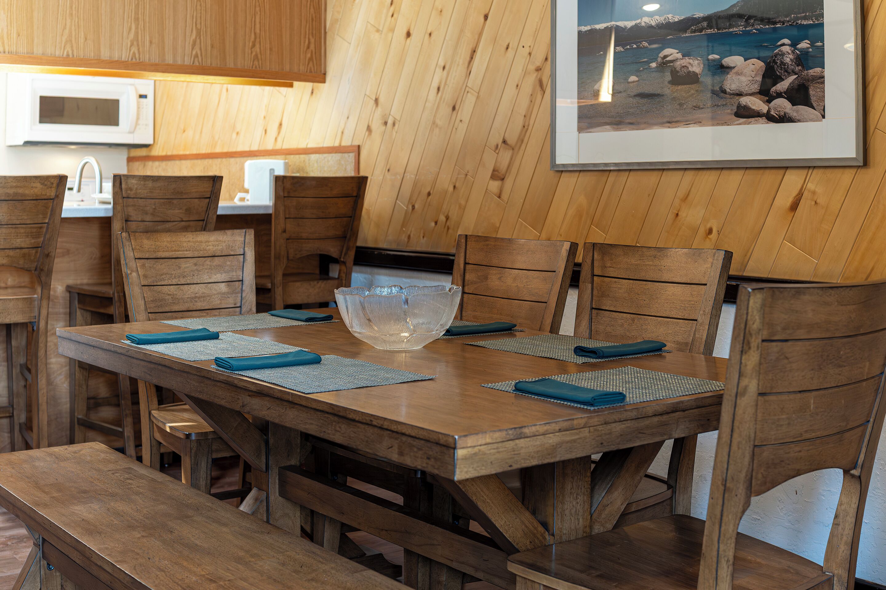 A wooden dining table with a large glass bowl centerpiece, surrounded by wooden chairs with blue placemats.
