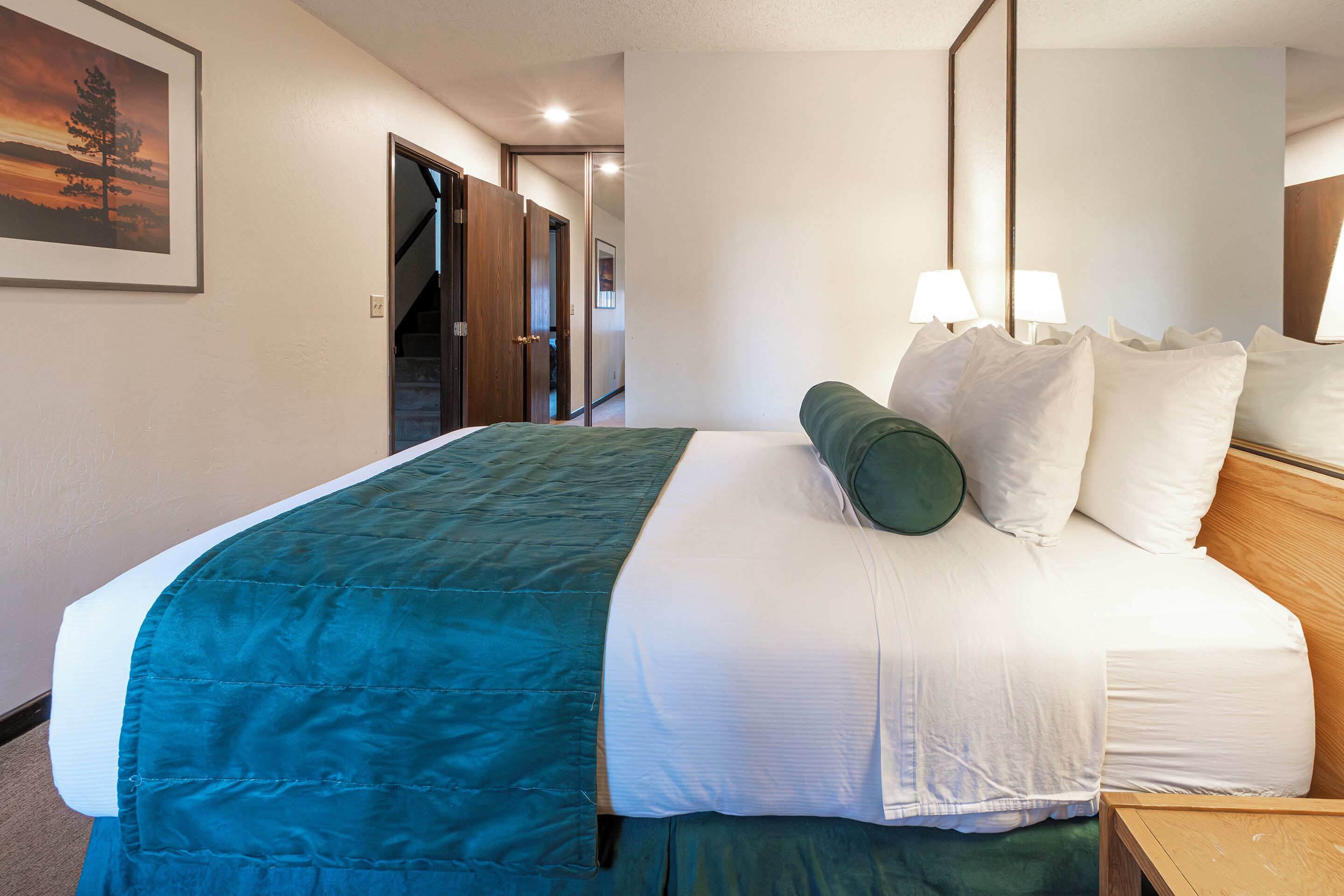 Un lit bien fait, recouvert d’une couvre-lit turquoise et blanche, avec deux oreillers blancs et un oreiller vert. Une table de chevet en bois avec une lampe se trouve à côté du lit.