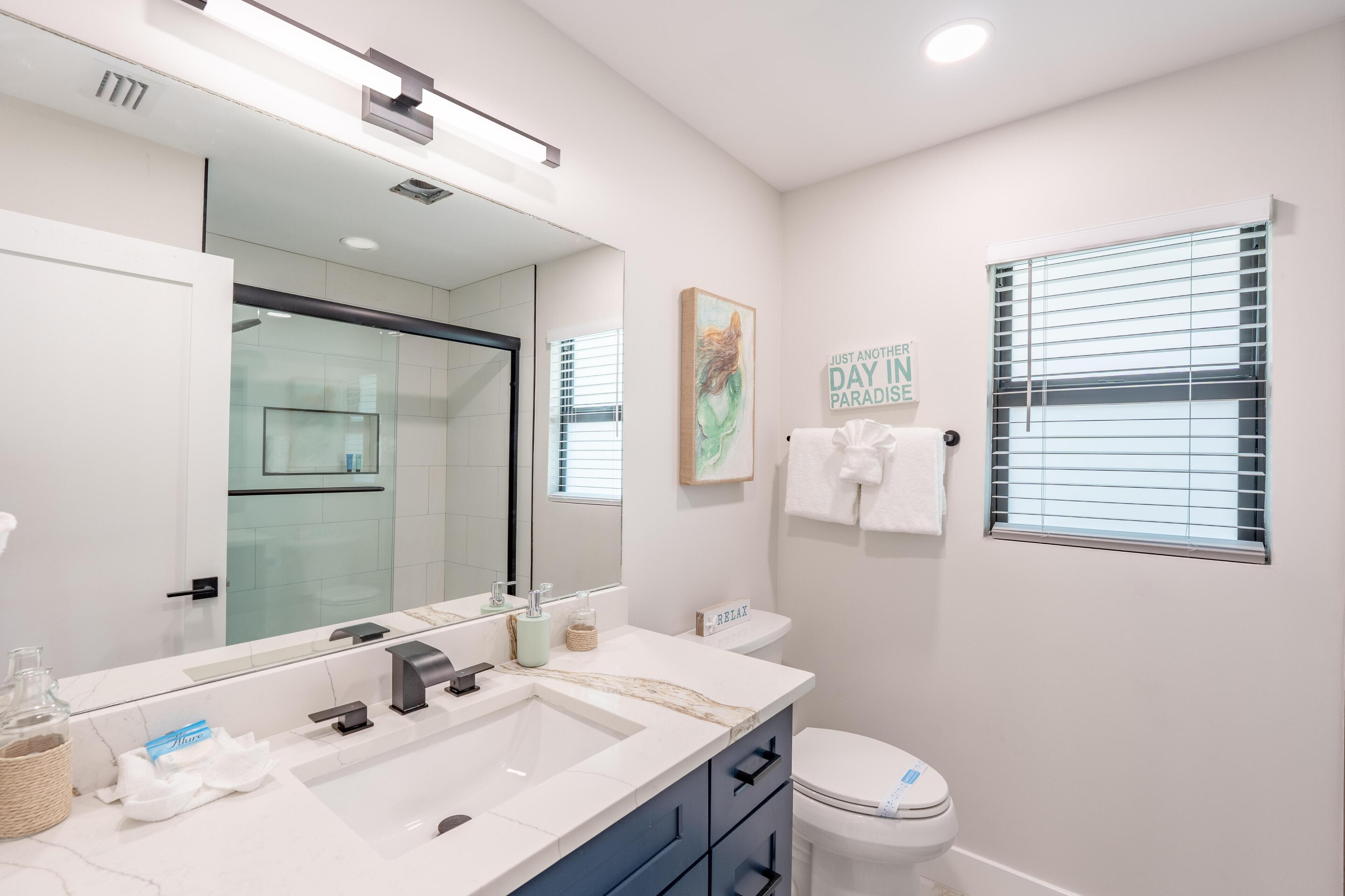 Une salle de bain moderne avec un grand miroir, un lavabo blanc, des toilettes et une fenêtre équipée de stores.