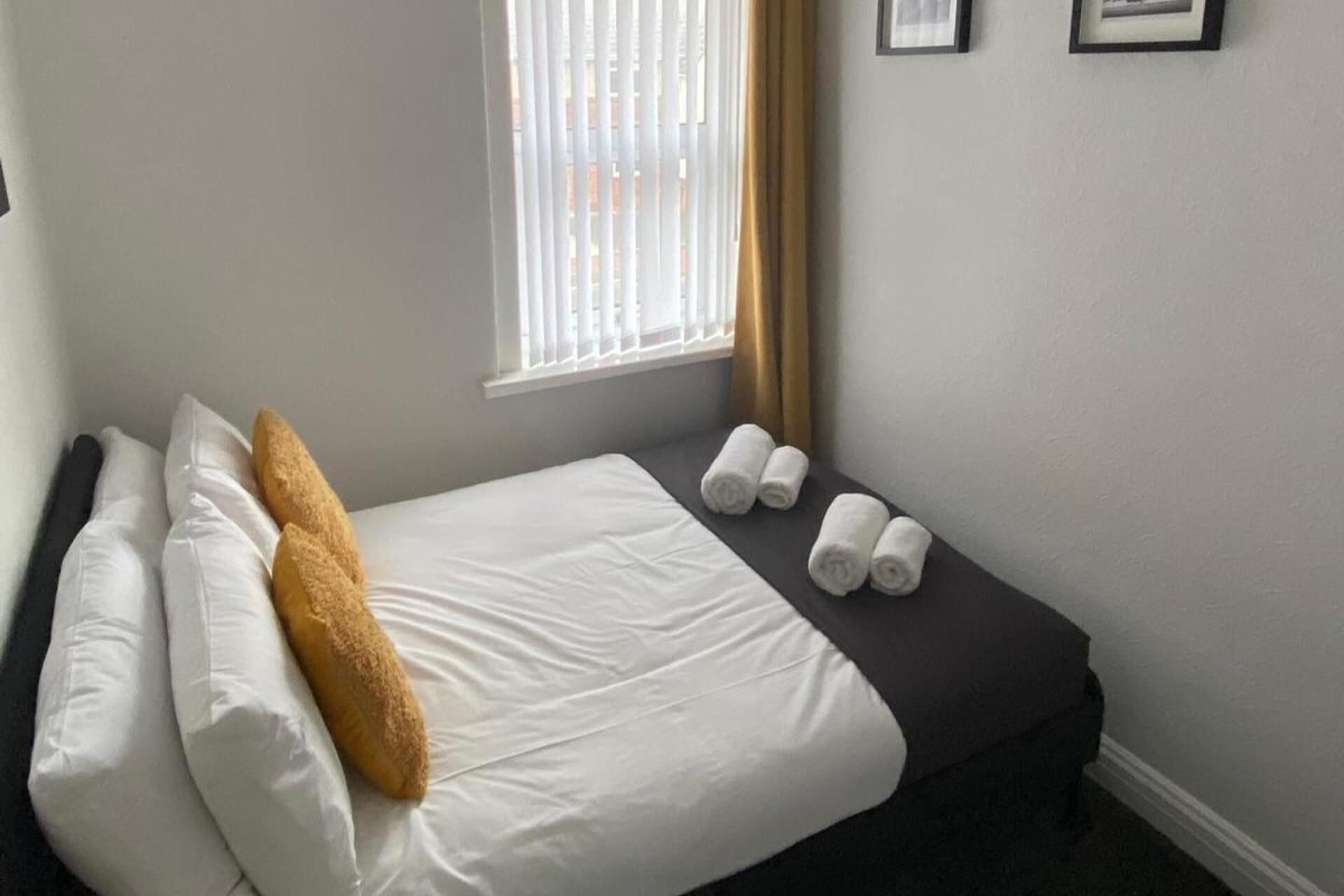 A hotel room with a bed, pillows, a window with blinds, and rolled towels on the bed.