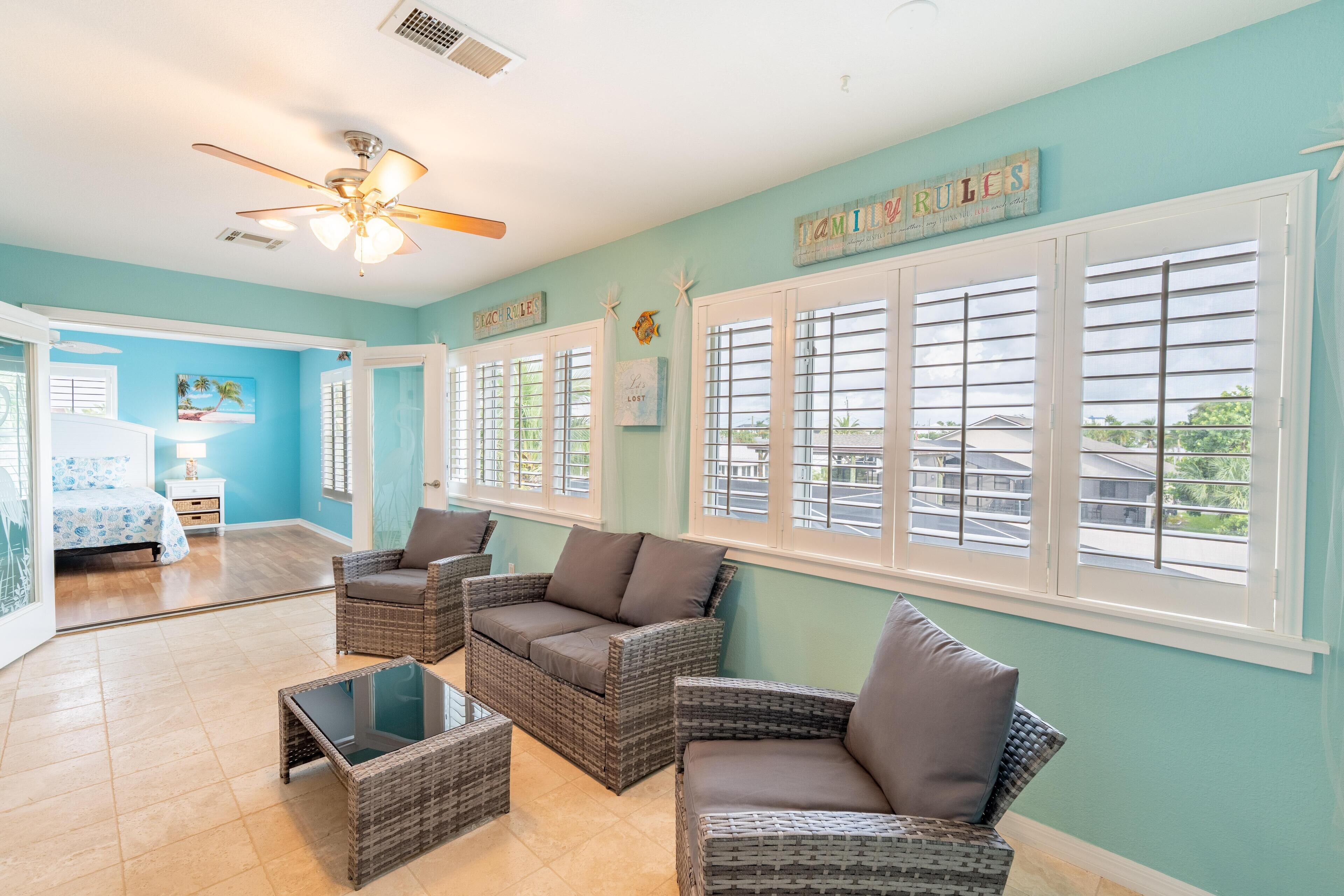 A room with wicker furniture, a ceiling fan, and large windows with shutters.