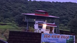 A villa with a red roof and white walls, surrounded by lush greenery and hills. A signboard in front advertises the villa as family-friendly with amenities like free Wi-Fi, parking, and smart TV. - Sea view villa (Shrivardhan)