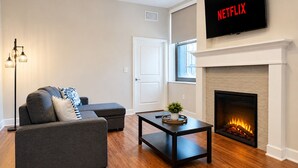 A living room with a sofa, a coffee table, a fireplace, and a TV mounted above the fireplace. - Cozy Luxe Suite R&R HoF, Casino, Stadium, Airport (Cleveland)