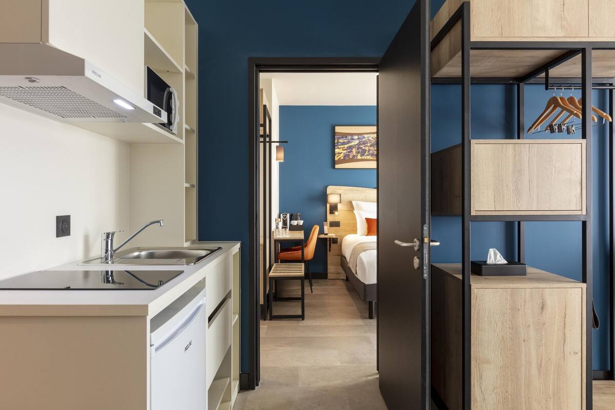 A compact hotel room with a kitchenette, a bed, a desk, and a wardrobe.