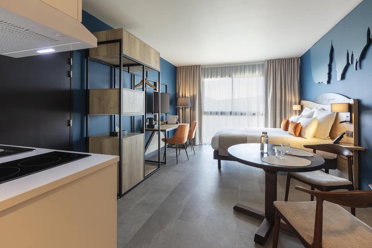 A modern hotel room with a bed, a desk, and a kitchenette.