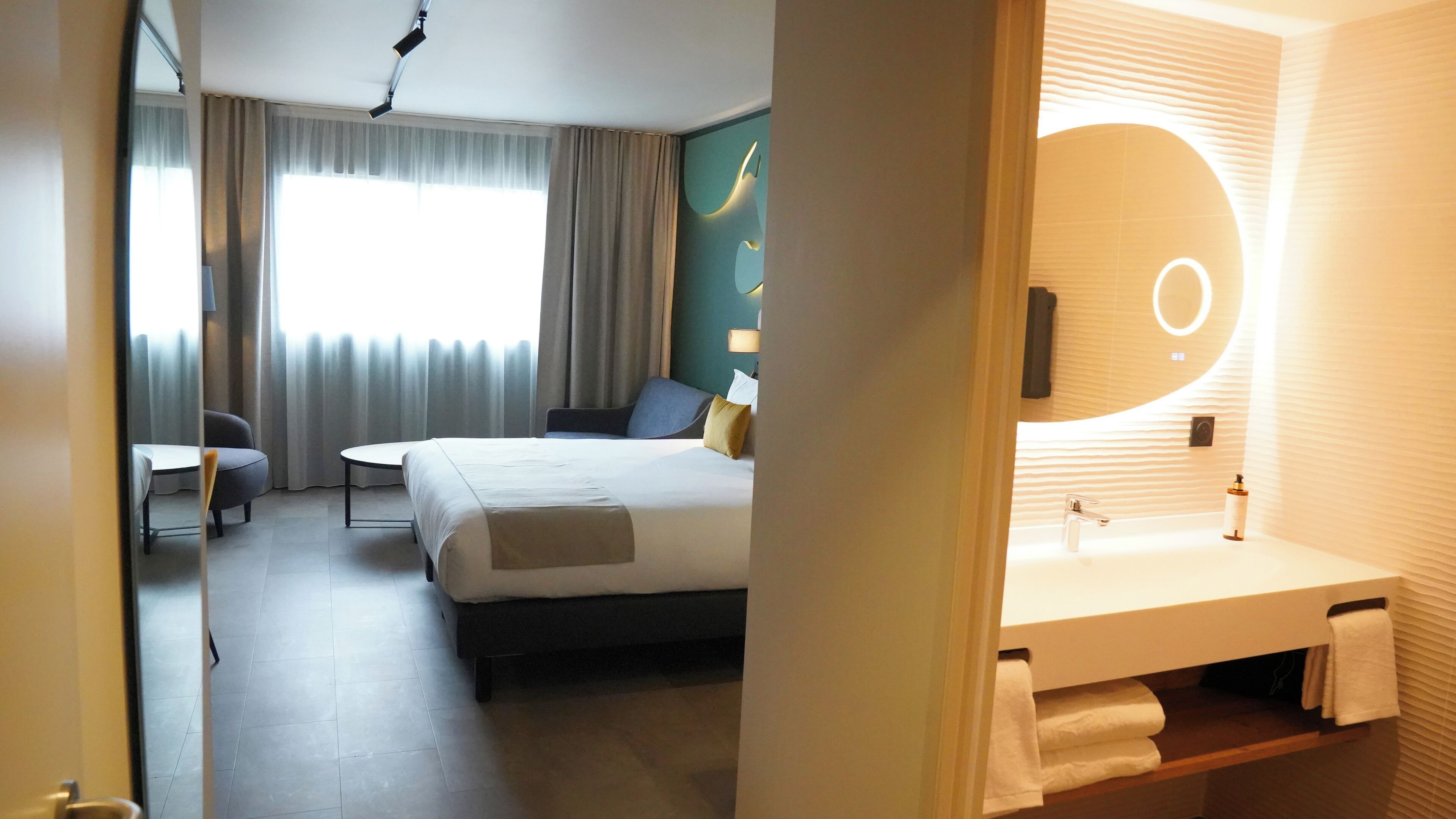 A hotel room with a bed, a desk, a chair, and a bathroom with a sink and mirror.