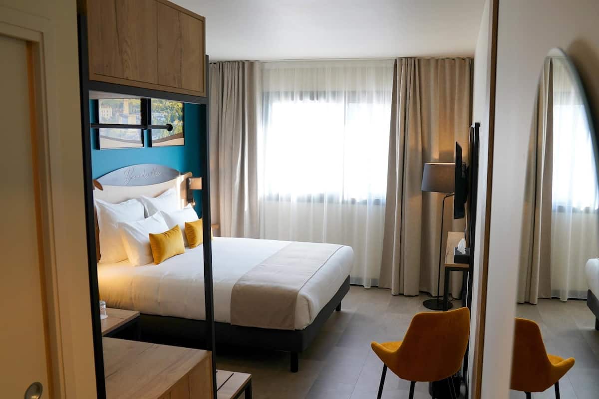 A hotel room with a bed, a desk, two chairs, a TV, and a window with curtains.