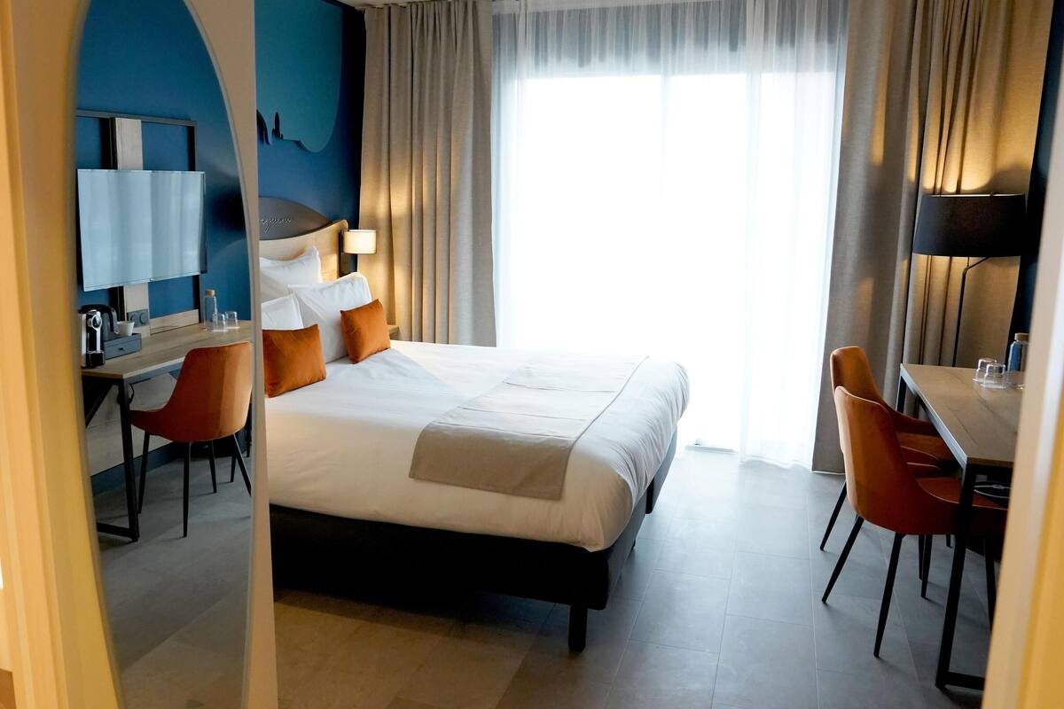 A hotel room with a bed, a desk, a chair, a mirror, and a window with curtains.