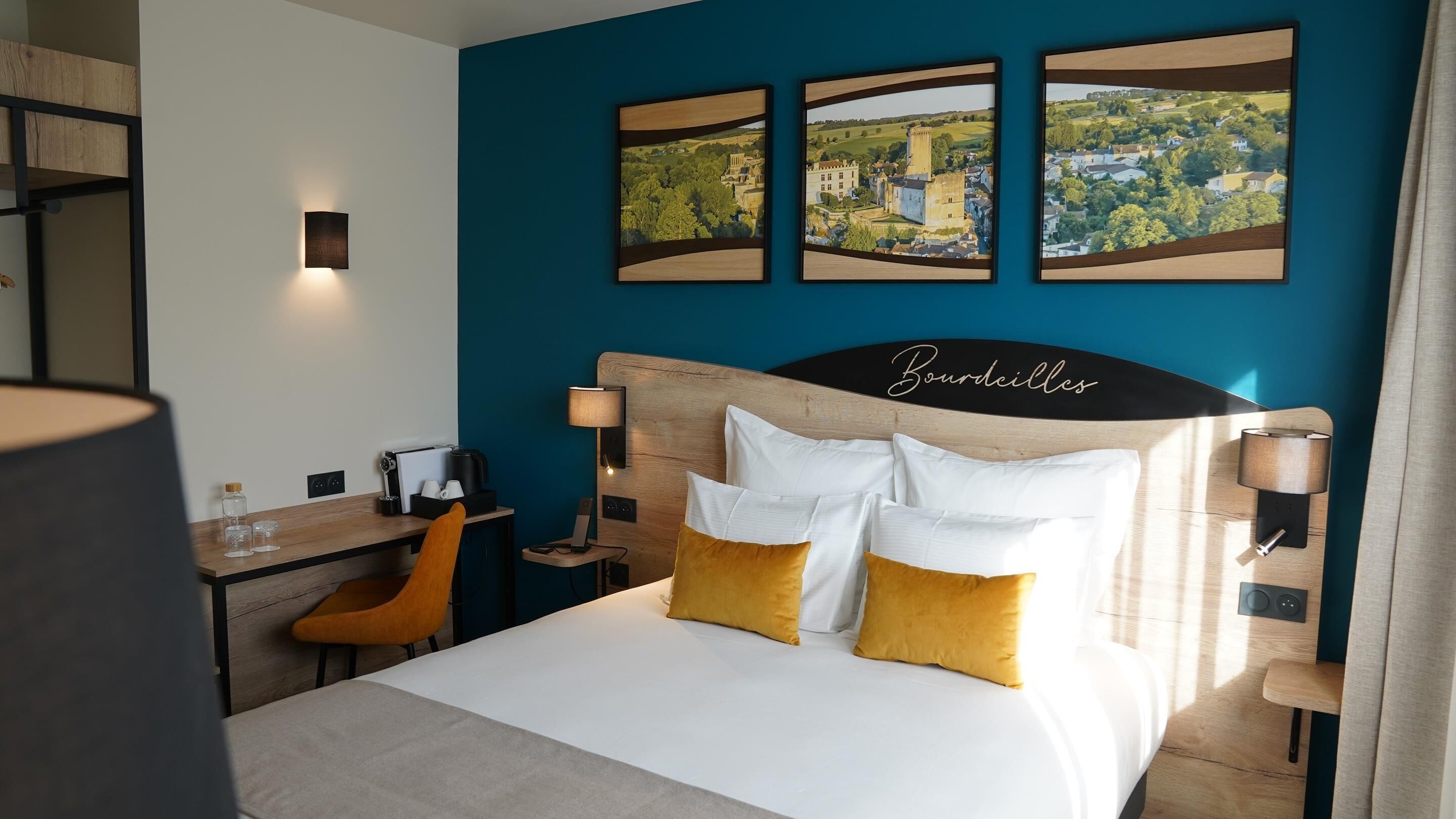 A hotel room with a bed, a desk, a chair, and three framed pictures on the wall.