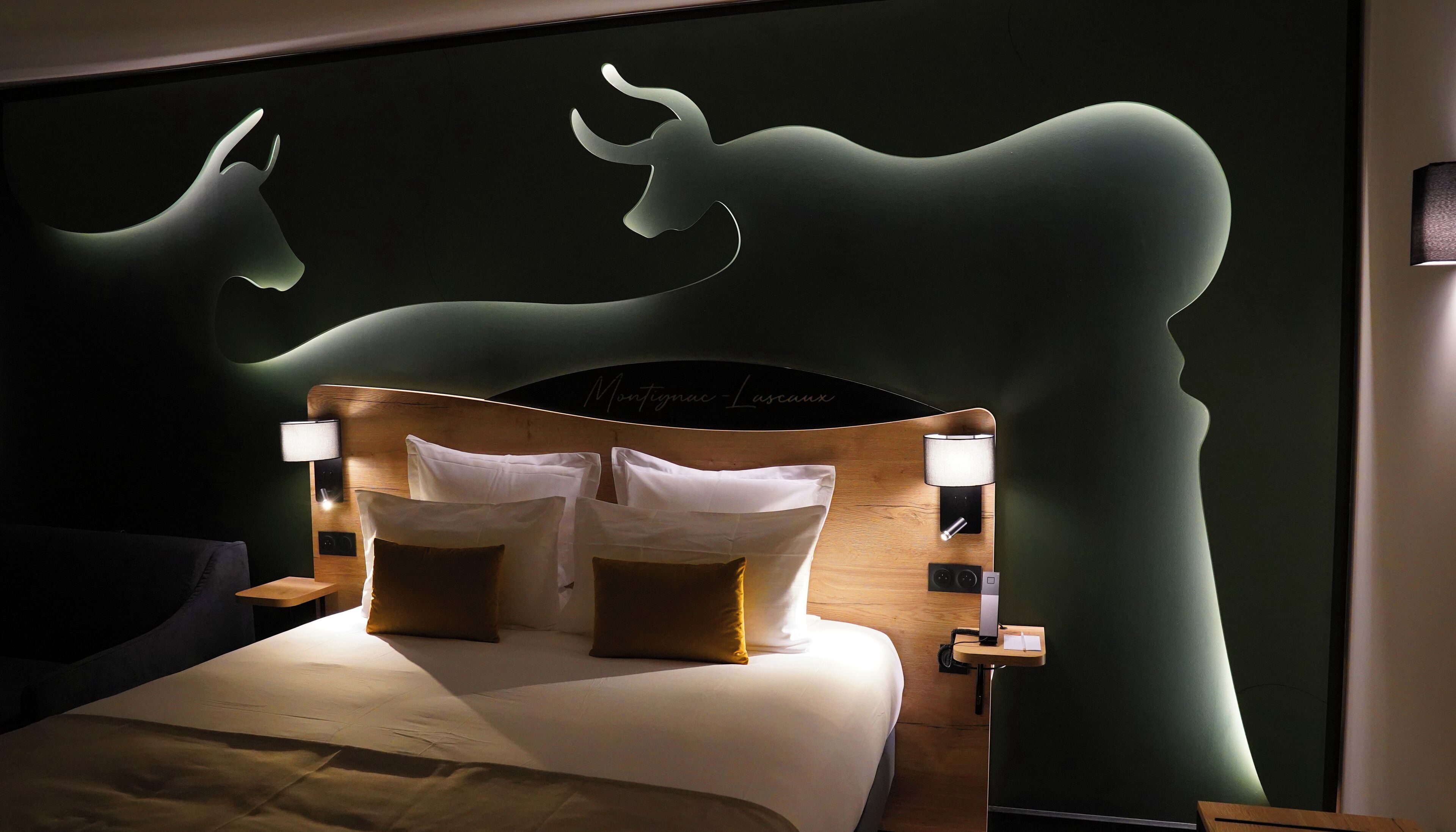 A bed with white linens, two brown pillows, and a wooden headboard with a silhouette of a deer. There are two bedside lamps on either side of the bed.