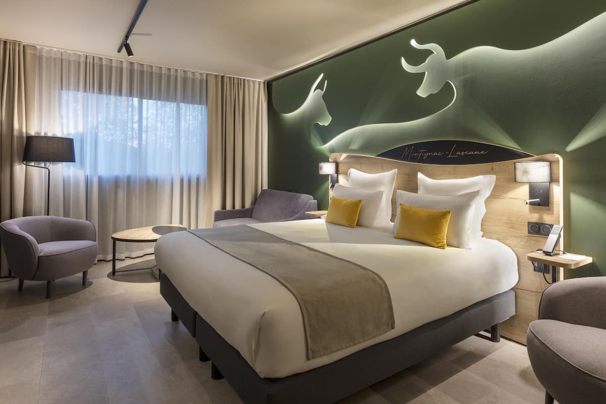 A modern hotel room with a large bed, two armchairs, a small table, and a wall mural.