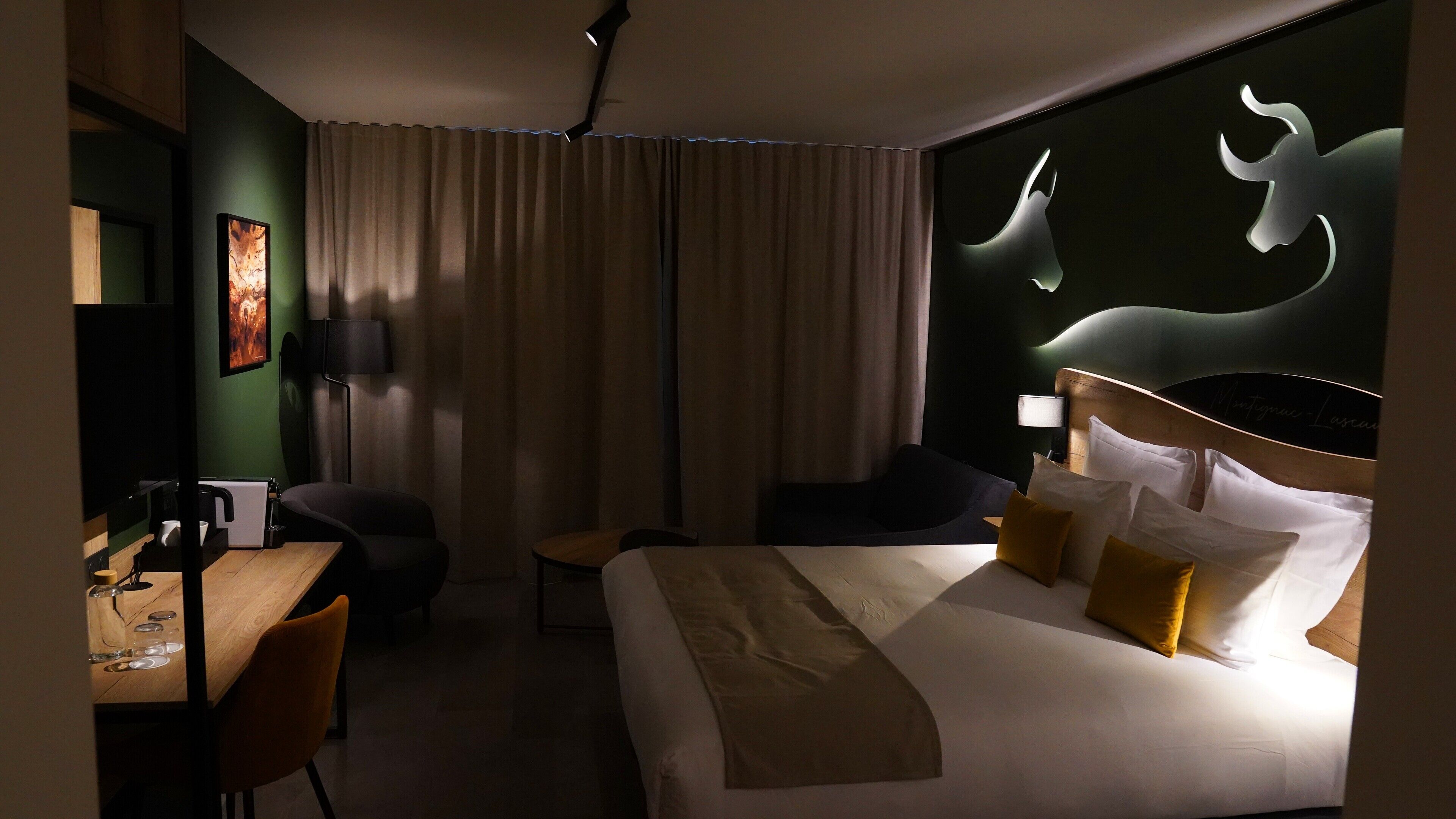 Ein Hotelzimmer mit Bett, zwei Stühlen, einem Schreibtisch mit Lampe, einem großen Fenster mit Vorhängen und einer Wand mit einer Lichtskulptur.