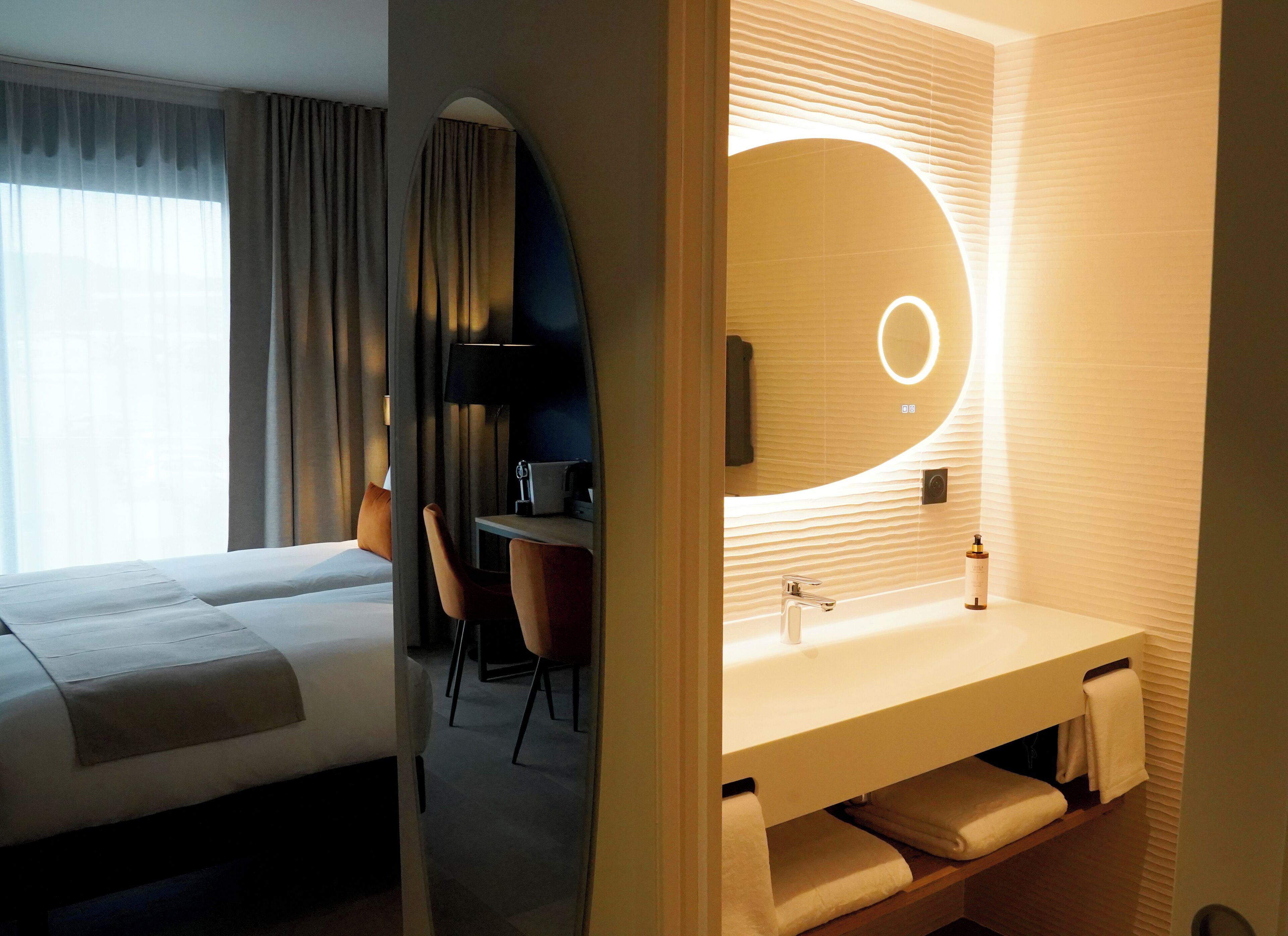 Ein Hotelzimmer mit zwei Betten, einem Schreibtisch und einem modernen Badezimmer mit großem Spiegel und integriertem Waschbecken.