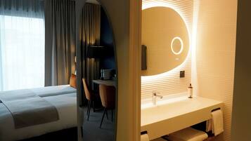 Ein Hotelzimmer mit zwei Betten, einem Schreibtisch und einem modernen Badezimmer mit großem Spiegel und integriertem Waschbecken.