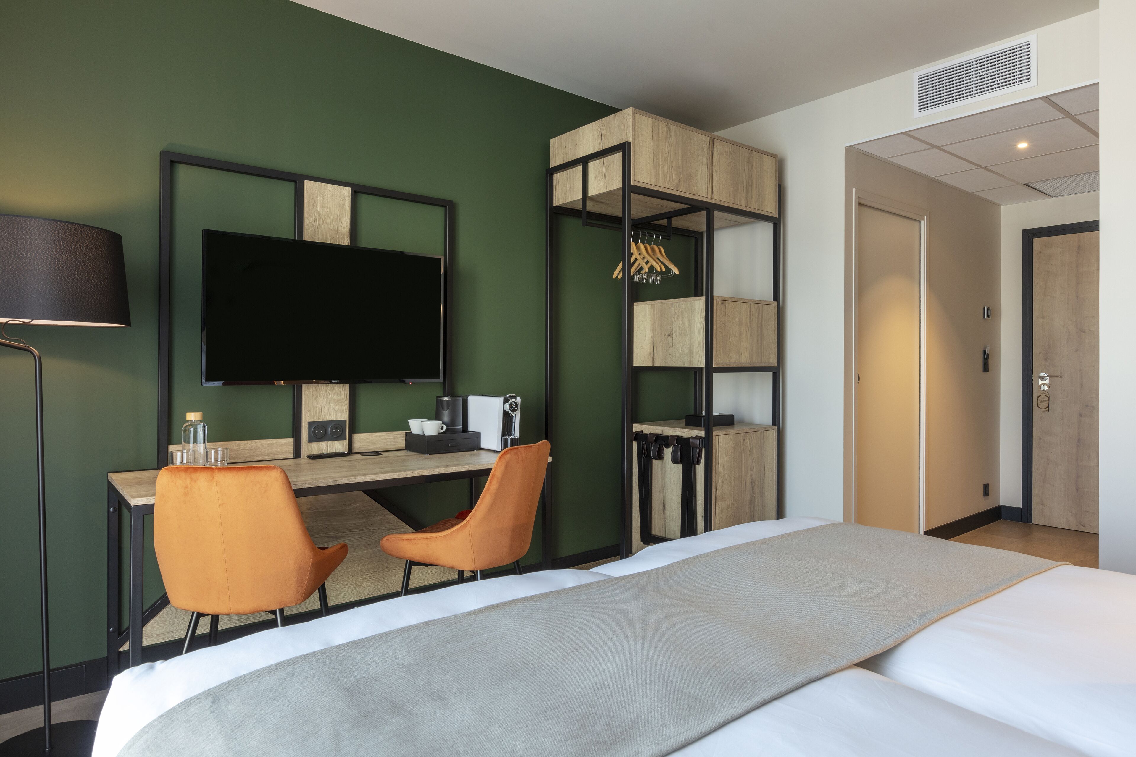 Une chambre d’hôtel comprenant un lit, deux chaises, un bureau avec un téléviseur à écran plat, une machine à café et une armoire avec des vêtements suspendus.