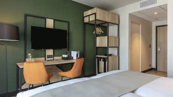 Une chambre d’hôtel comprenant un lit, deux chaises, un bureau avec un téléviseur à écran plat, une machine à café et une armoire avec des vêtements suspendus.