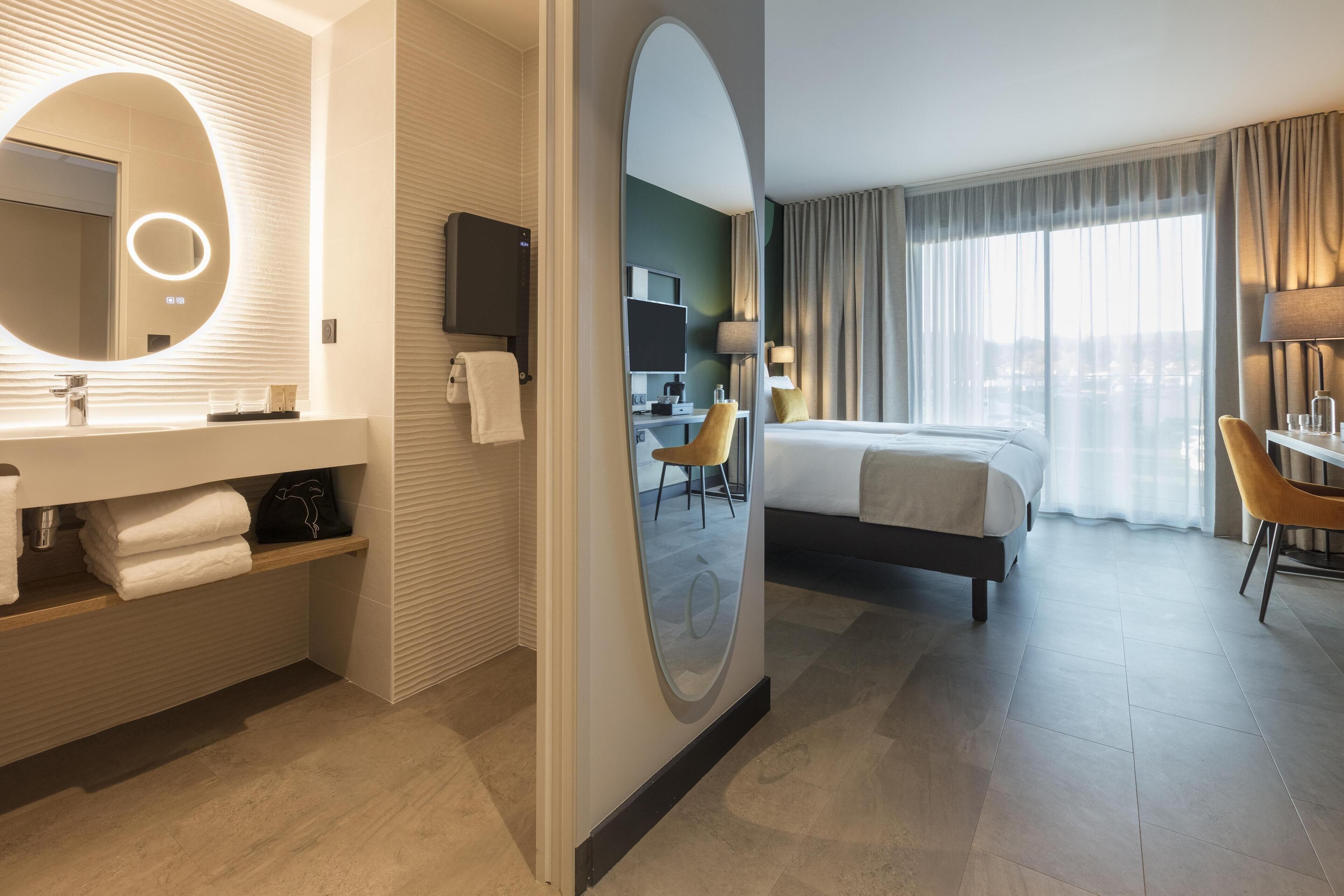 Una habitación de hotel moderna con una cama grande, un rincón de descanso con una silla, un escritorio, un televisor y un baño con espejo y lavamanos.