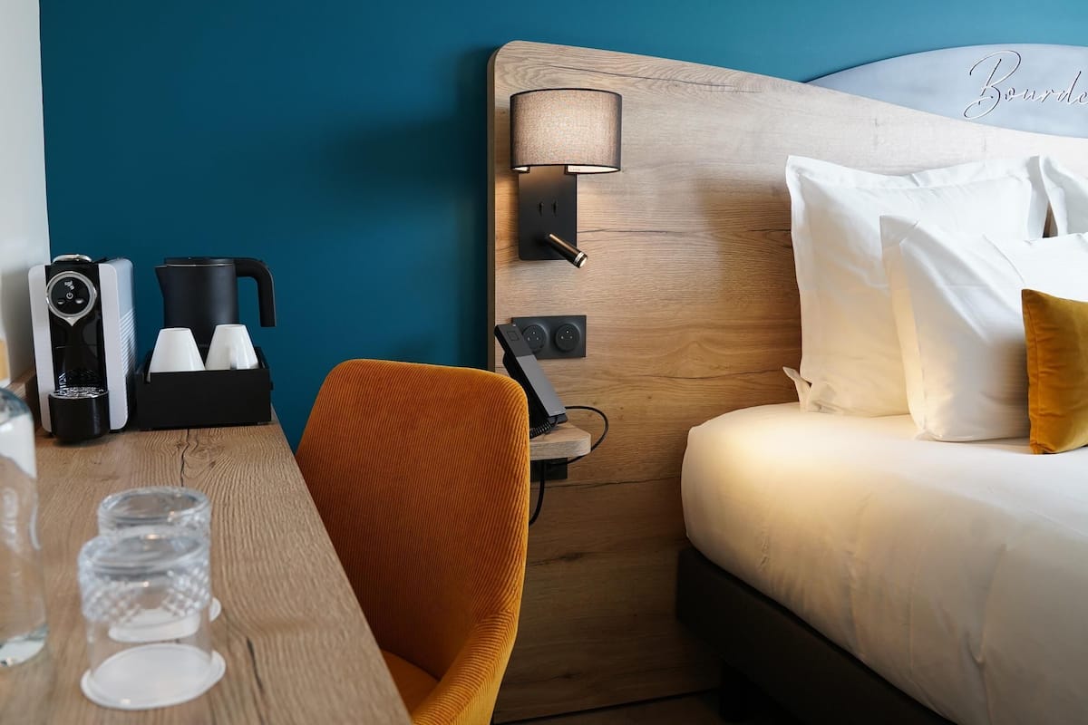 A hotel room with a wooden headboard, a coffee maker, a glass of water, and a bed with white bedding.