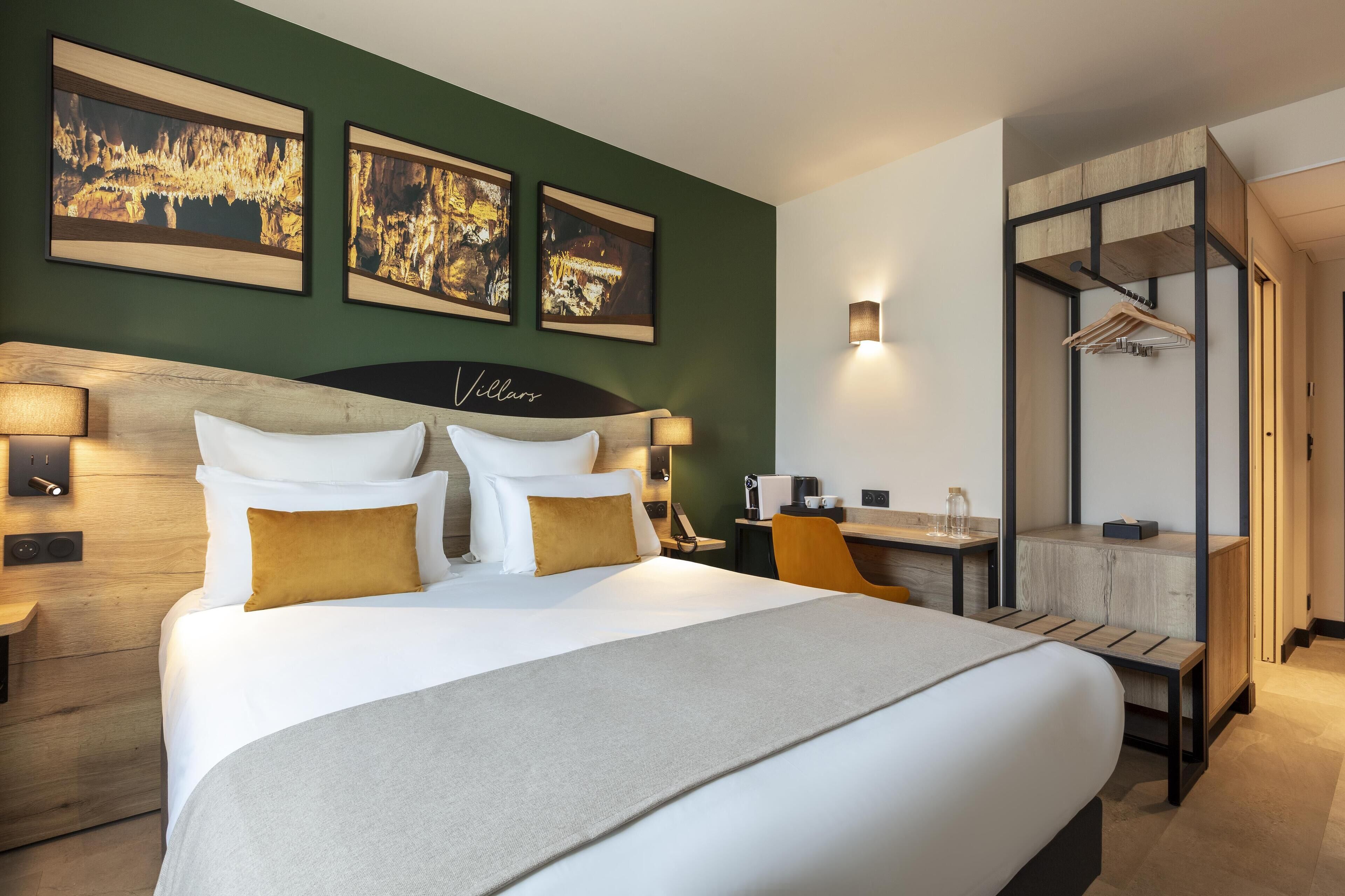Une chambre d’hôtel avec un grand lit, un bureau et une chaise. Trois tableaux encadrés sont accrochés au mur, au-dessus du lit.