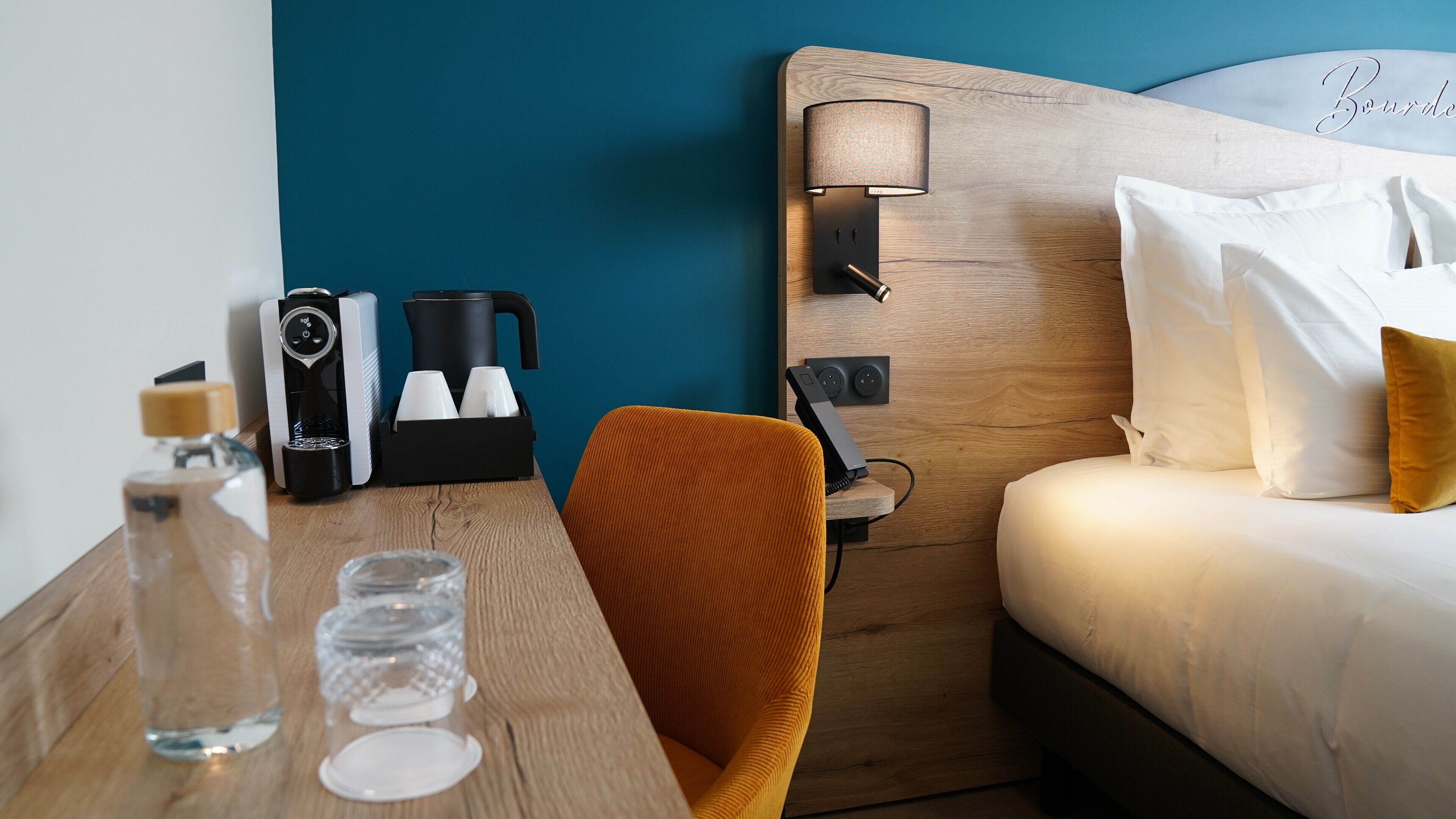 Una habitación de hotel con cabecera de madera, cafetera, teléfono y cama con ropa blanca.
