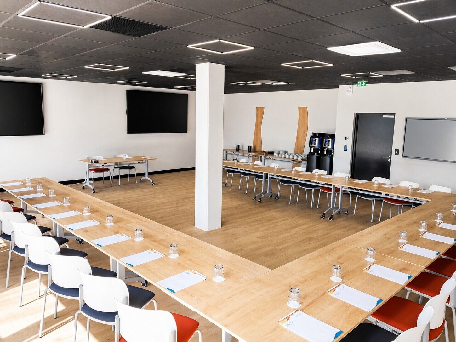 A conference room with long tables, chairs, a projector screen, and a coffee machine.