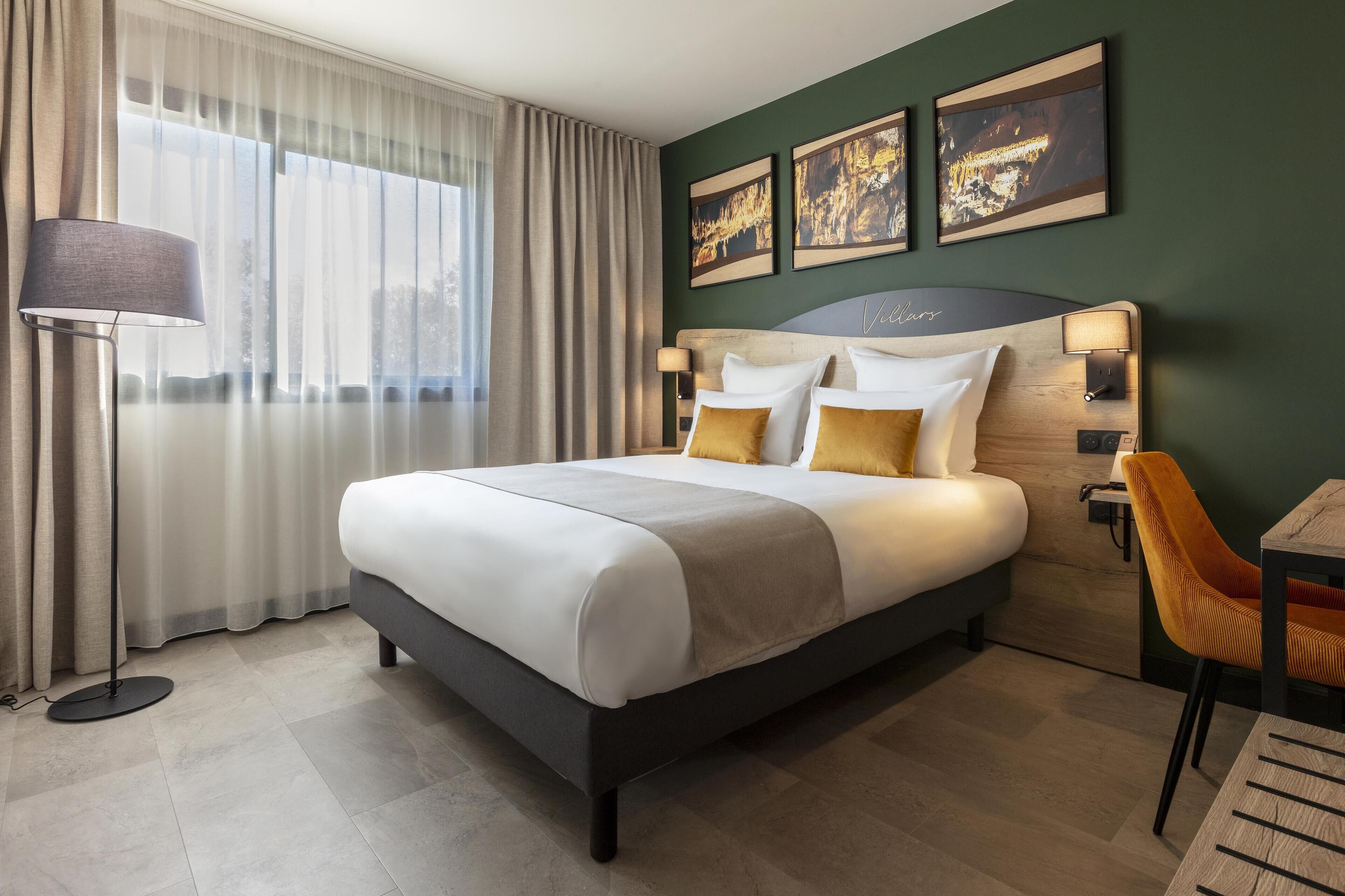 A modern hotel room with a large bed, a desk, and a chair. There are three framed pictures on the wall, and a floor lamp is visible.