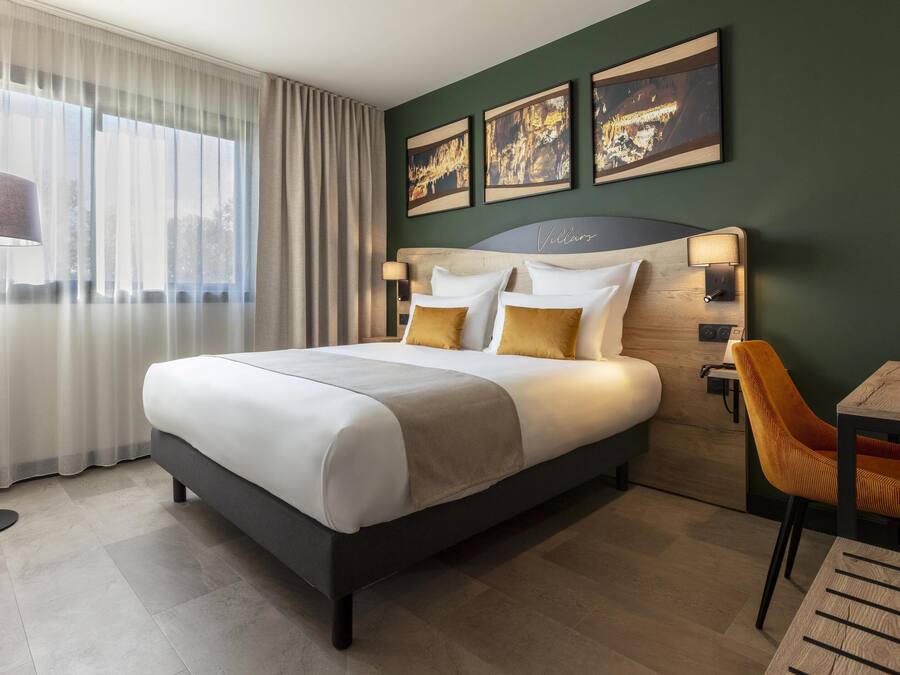 A modern hotel room with a large bed, a desk, and a chair. There are three framed pictures on the wall, and a floor lamp is visible.