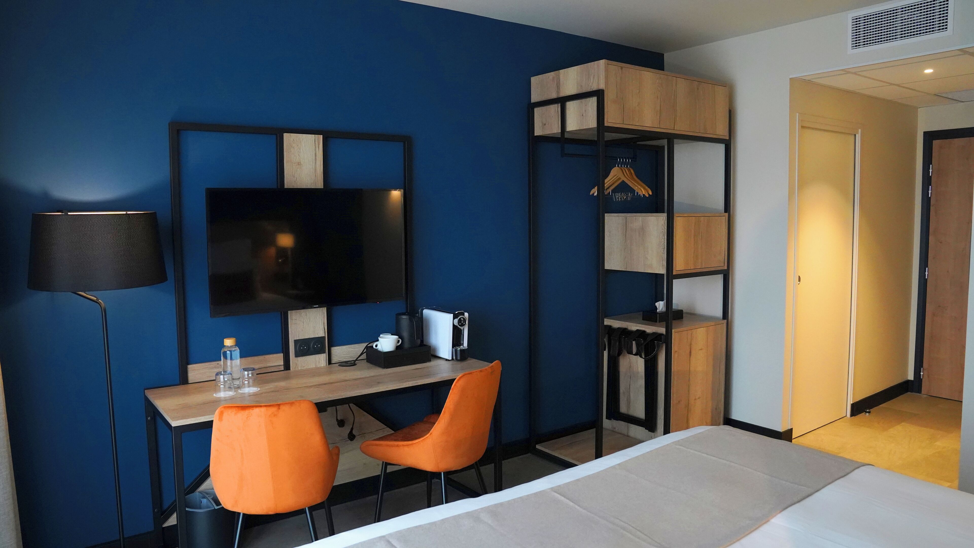 A hotel room with a bed, a desk with two orange chairs, a TV, and a wardrobe with a mirror.