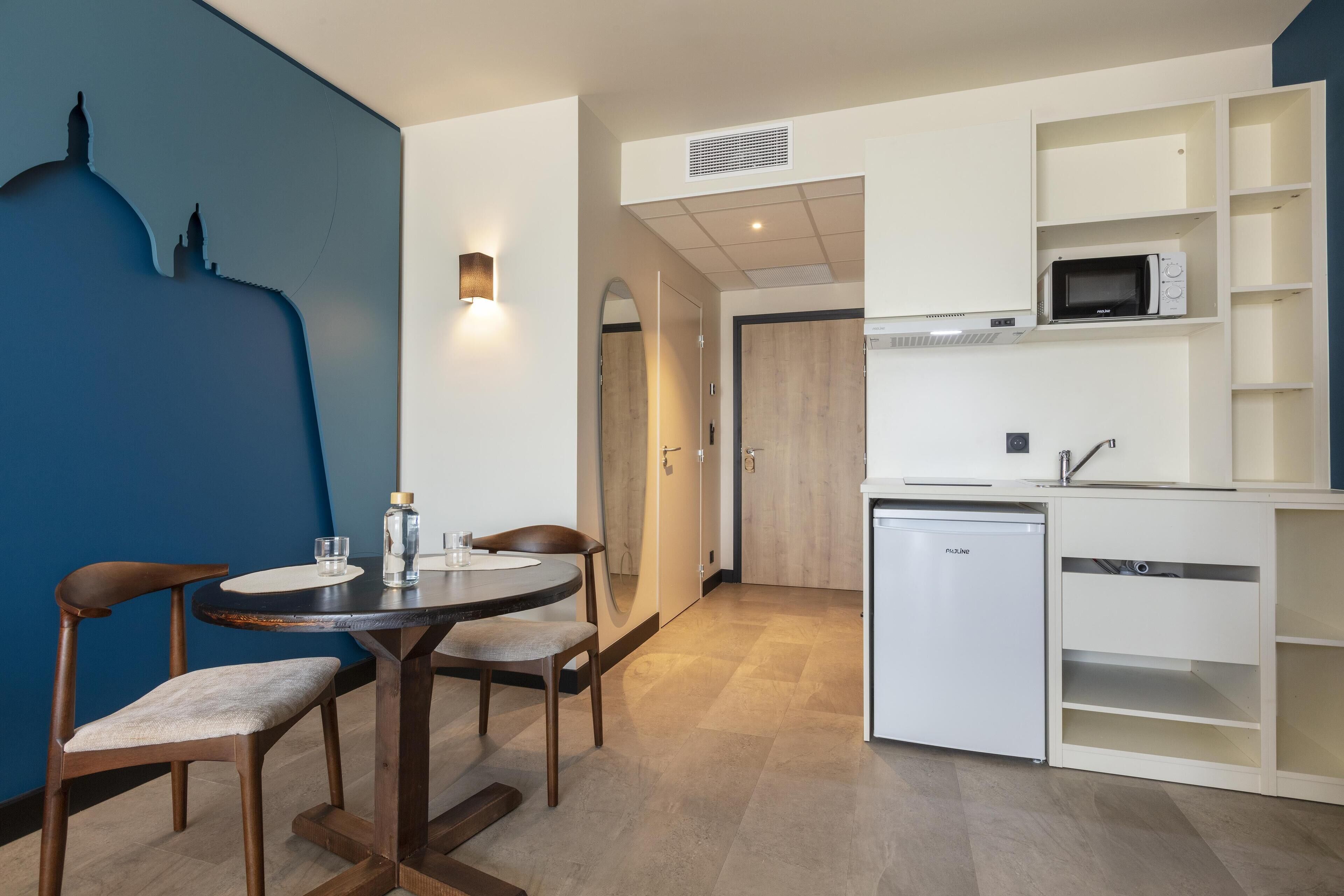 Une cuisine compacte équipée d’un four à micro-ondes, d’un lave-vaisselle et d’un meuble blanc. On y trouve une table à manger avec des chaises et un luminaire fixé au mur.