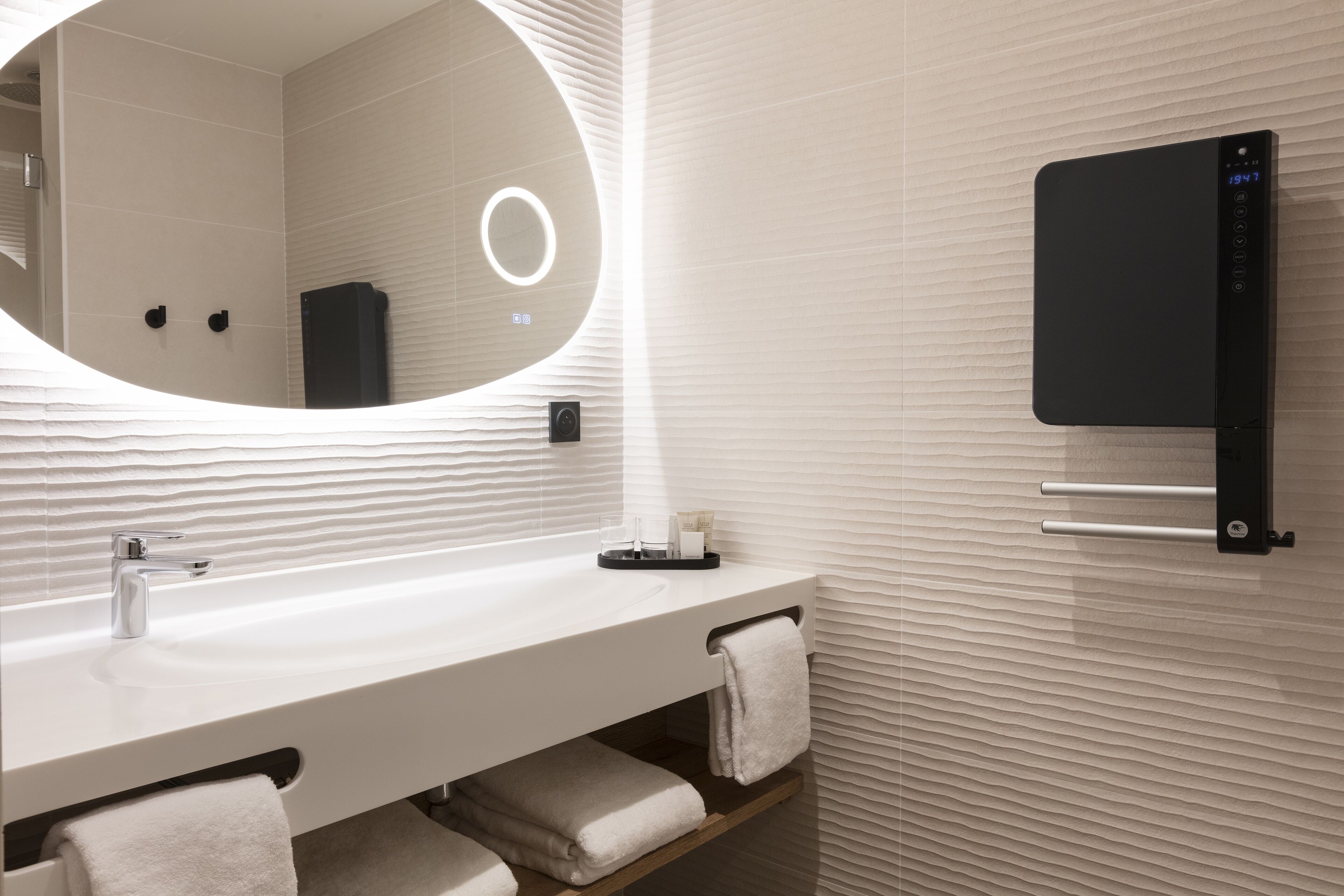 A modern bathroom with a white sink, a mirror with a circular light, a towel rack, and a black digital device on the wall.