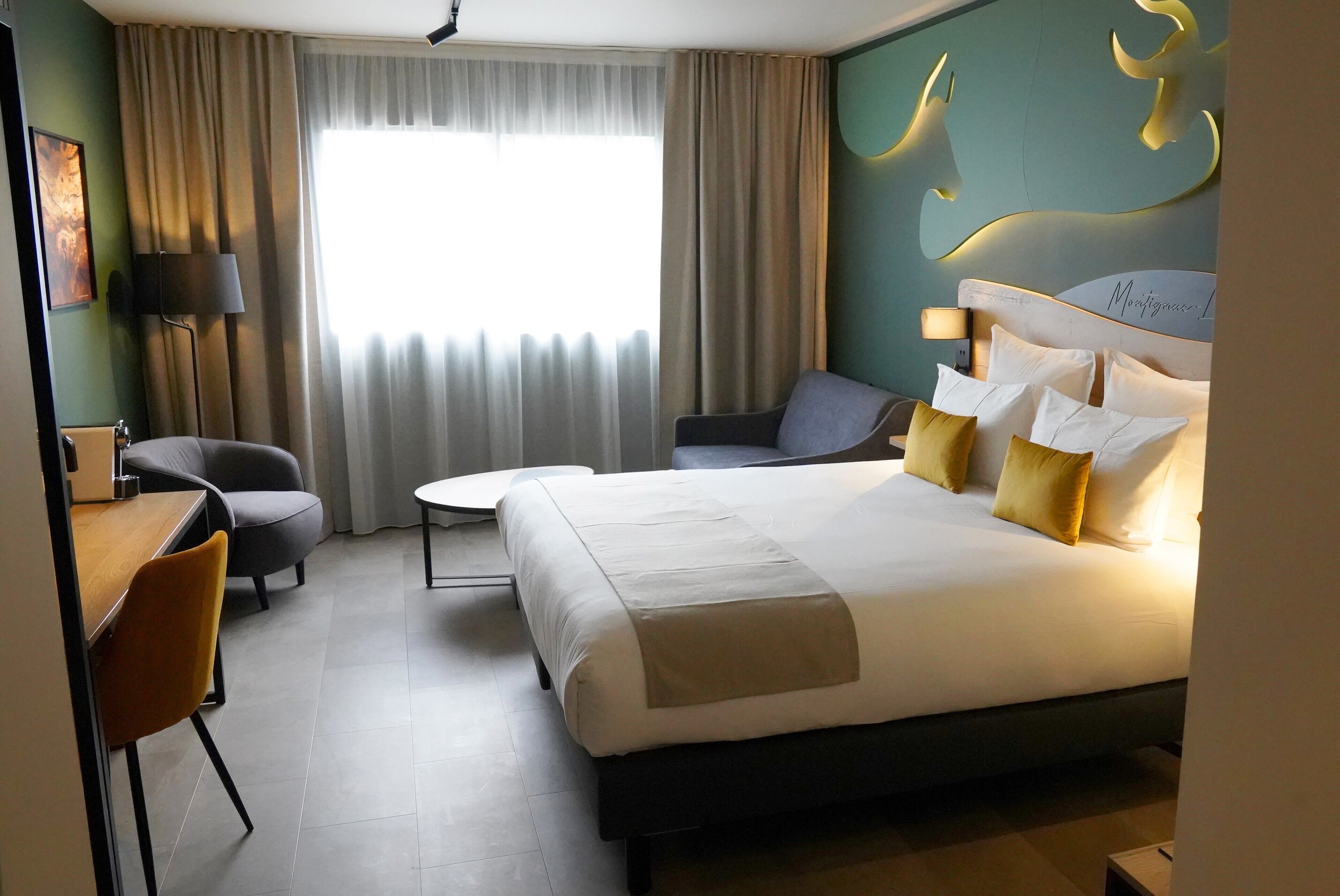A hotel room with a large bed, a desk, a chair, and a sofa. There is a window with curtains and a wall-mounted lamp.