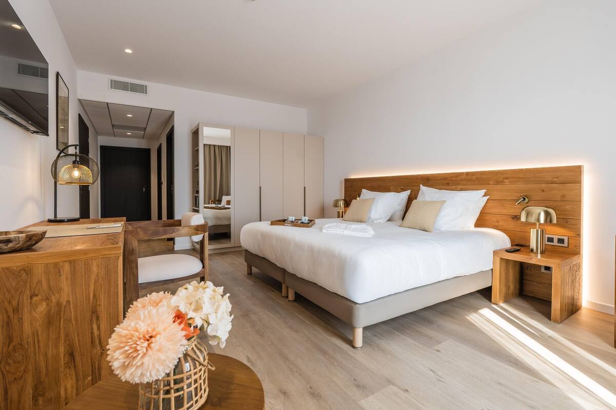 A modern hotel room with a large bed, wooden headboard, bedside tables, a desk, and a mirror.