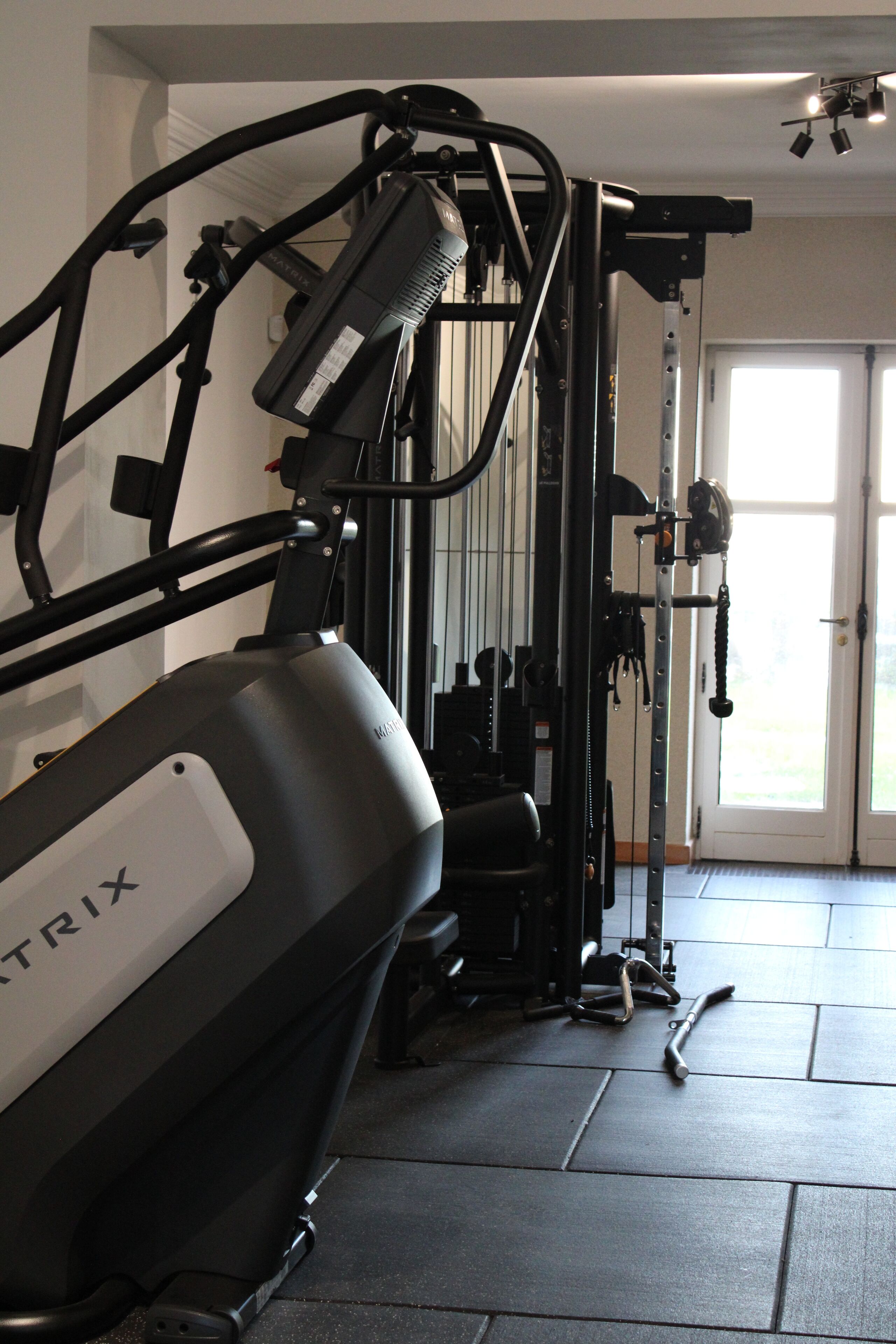 Un gimnasio en casa con una bicicleta estática, máquinas de pesas y una puerta que da a una habitación luminosa.