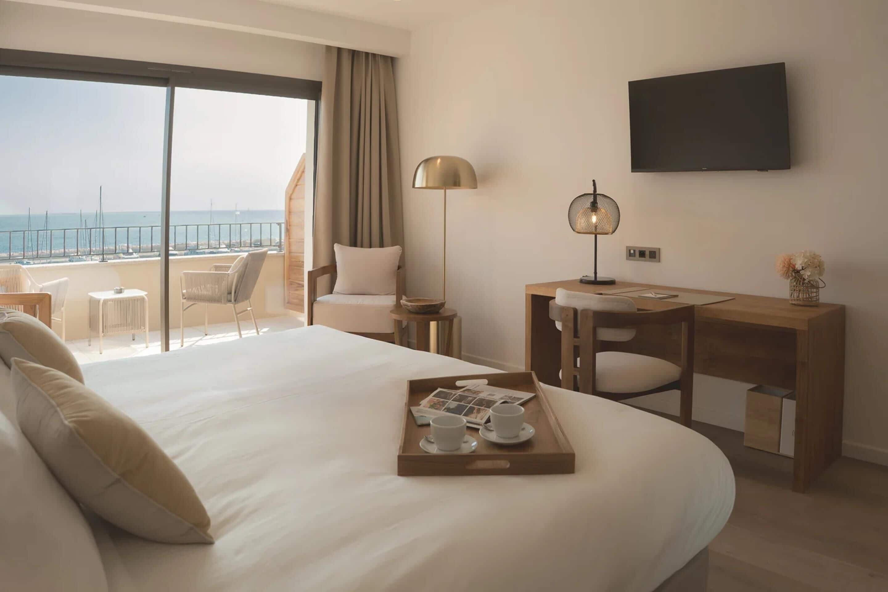 Een hotelkamer met een groot bed, een bureau, een stoel, een televisie en een balkon met uitzicht op zee.