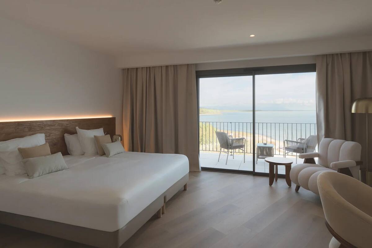 A hotel room with a large bed, a seating area with a table, and a view of the sea through a large window.